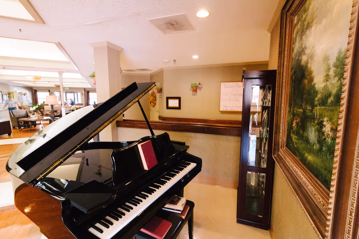 Interior view of a senior living facility common area featuring a black grand piano with sheet music on the stand. In the background, there are comfortable seating areas with chairs and tables, a large painting on the wall, and a glass display cabinet. The room is well-lit with ceiling lights and natural light from windows.
