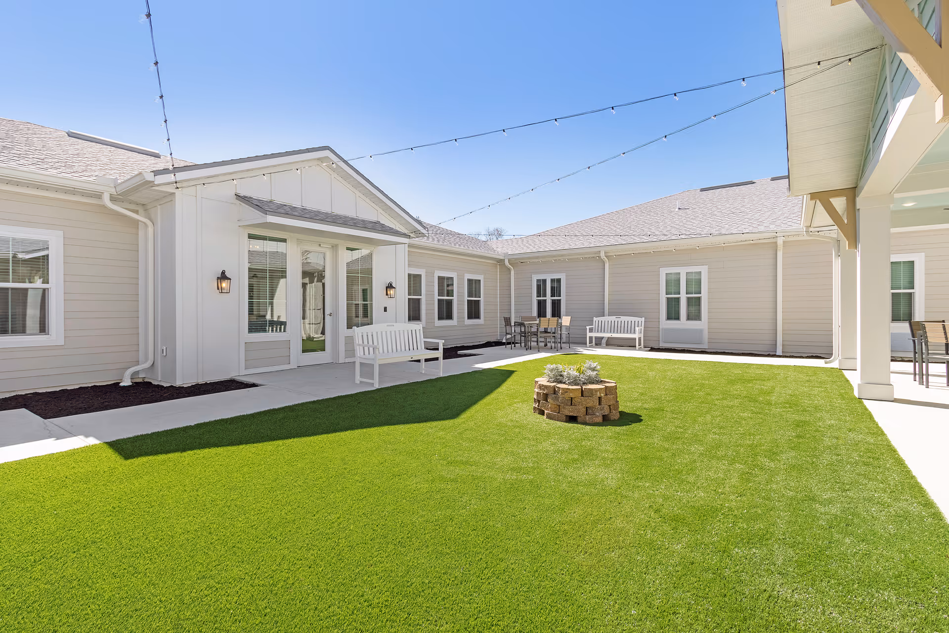 Outdoor courtyard area with artificial green grass, a small circular brick planter with plants in the center, white benches, patio tables with chairs, and string lights overhead. The surrounding building is light beige with white trim and multiple windows and doors.