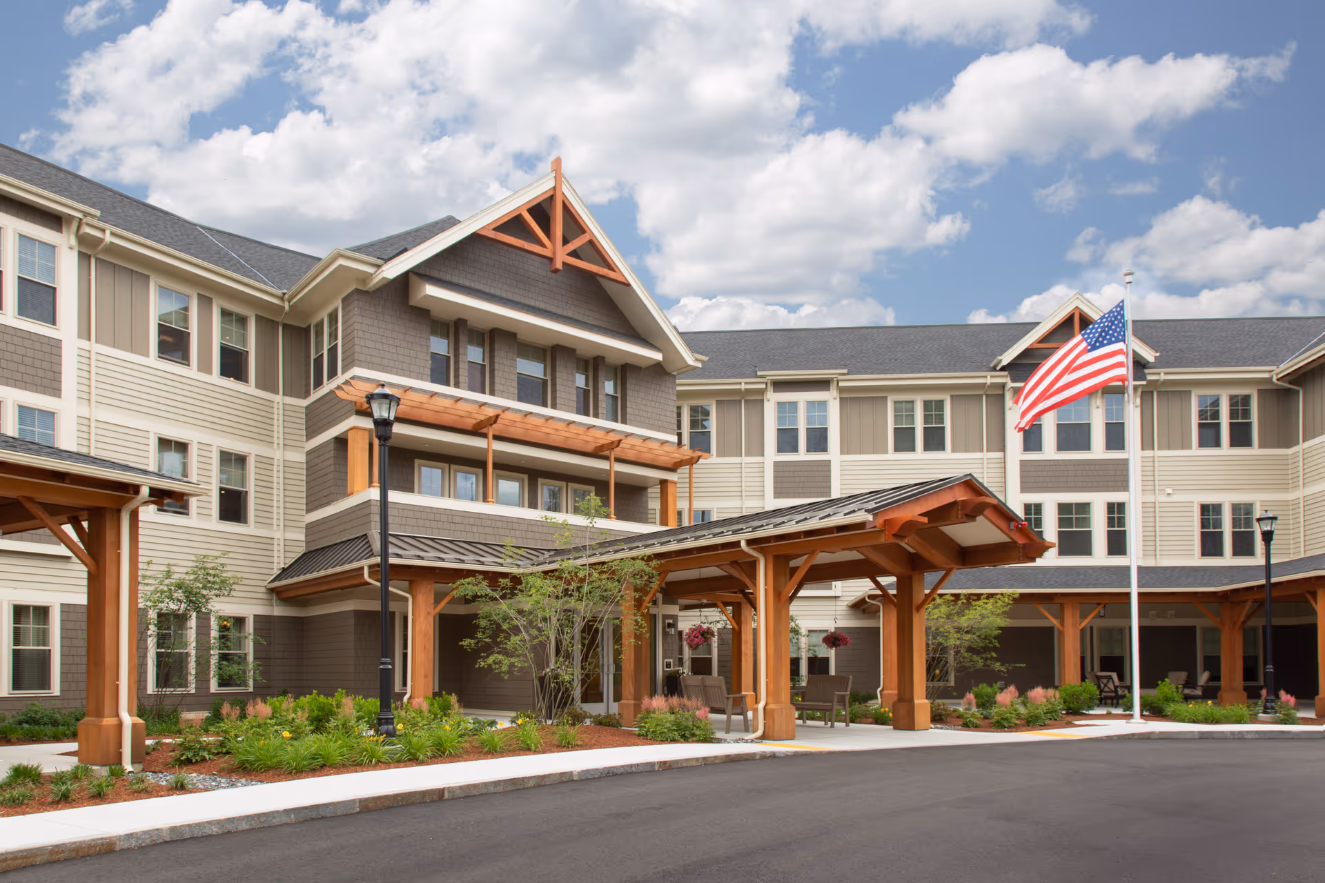 Front exterior view of a senior living facility named The Residence at Orchard Grove, featuring a three-story building with beige and brown siding, a covered entrance with wooden beams, landscaped greenery, and an American flag on a flagpole under a partly cloudy sky.