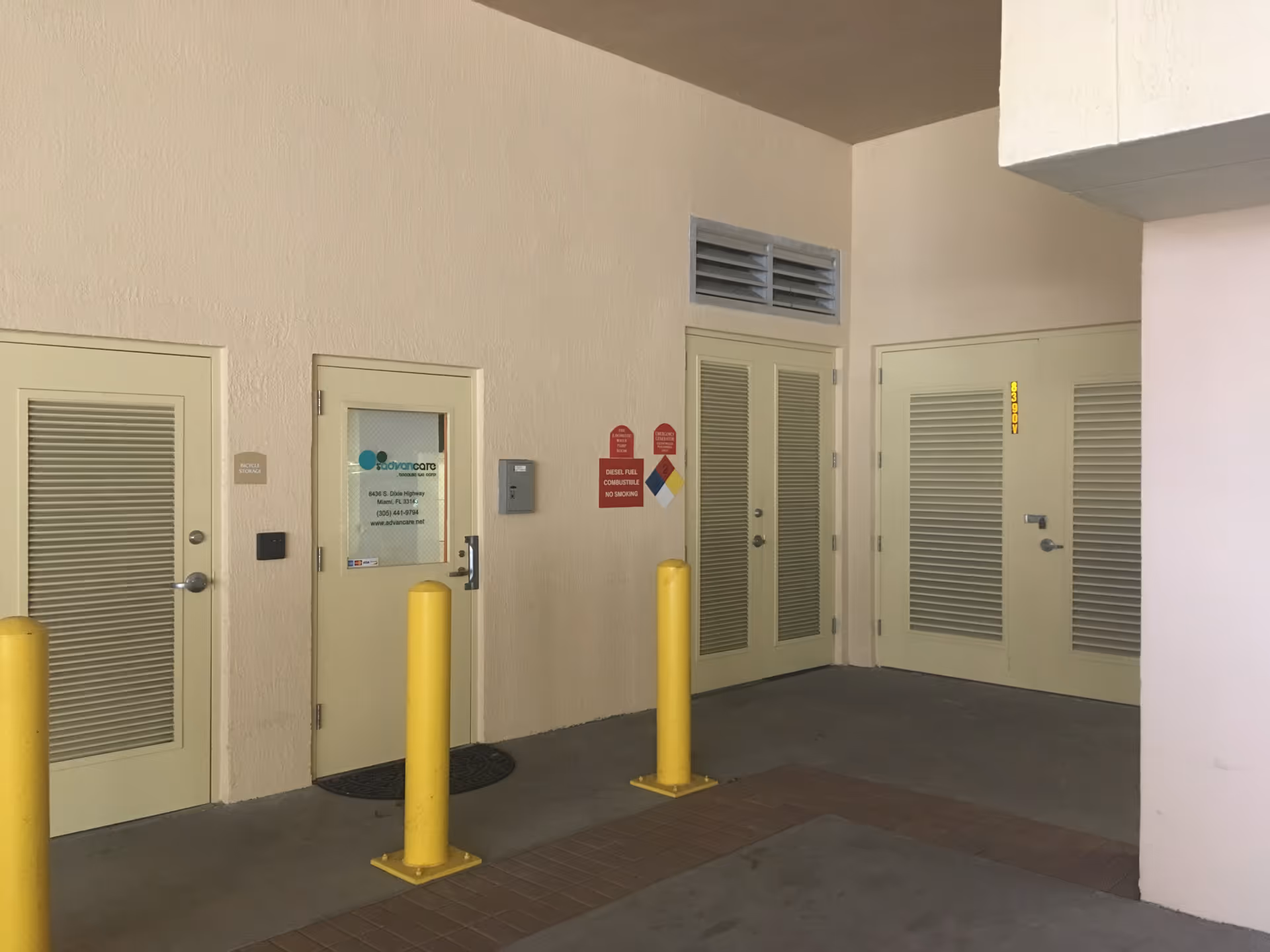 Beige indoor corridor with multiple louvered doors, yellow bollards, and signage.