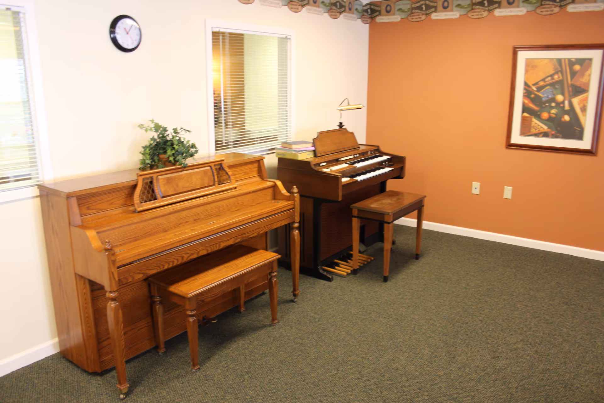 Wooden piano and organ sit in a cozy music room with orange walls and framed art.