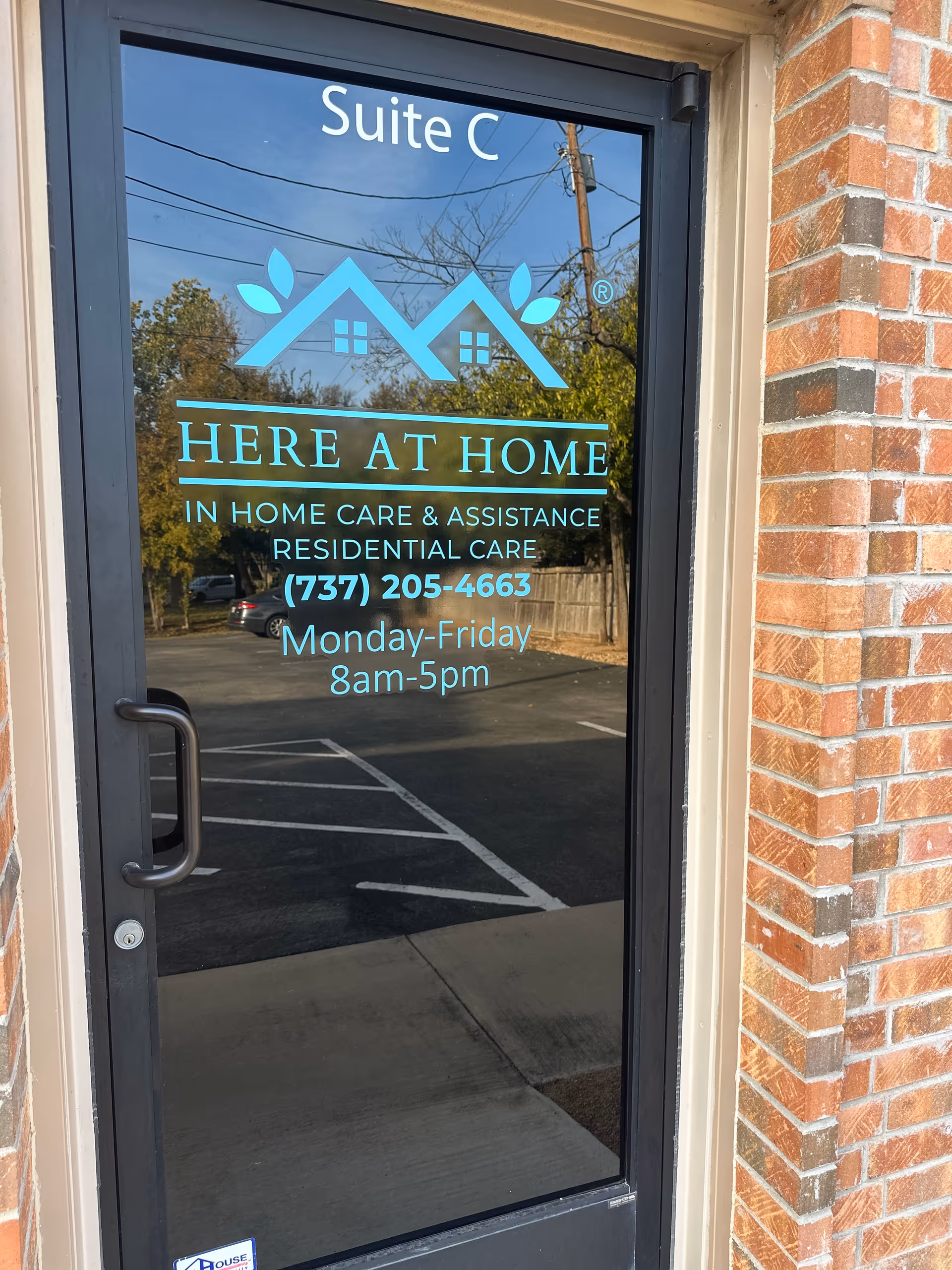 Glass door with blue HERE AT HOME sign, logo, and hours beside a brick building exterior.