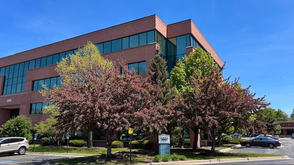 Brick building with large glass windows, surrounded by flowering trees and a parking lot.