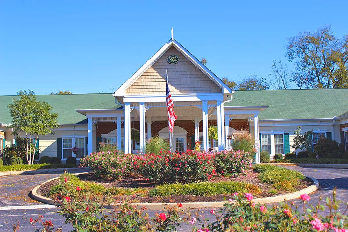 Front exterior view of Windsor Gardens Senior Living facility with a circular driveway, landscaped garden with blooming pink flowers, an American flag in the center, and a building with a green roof and white columns under a clear blue sky.