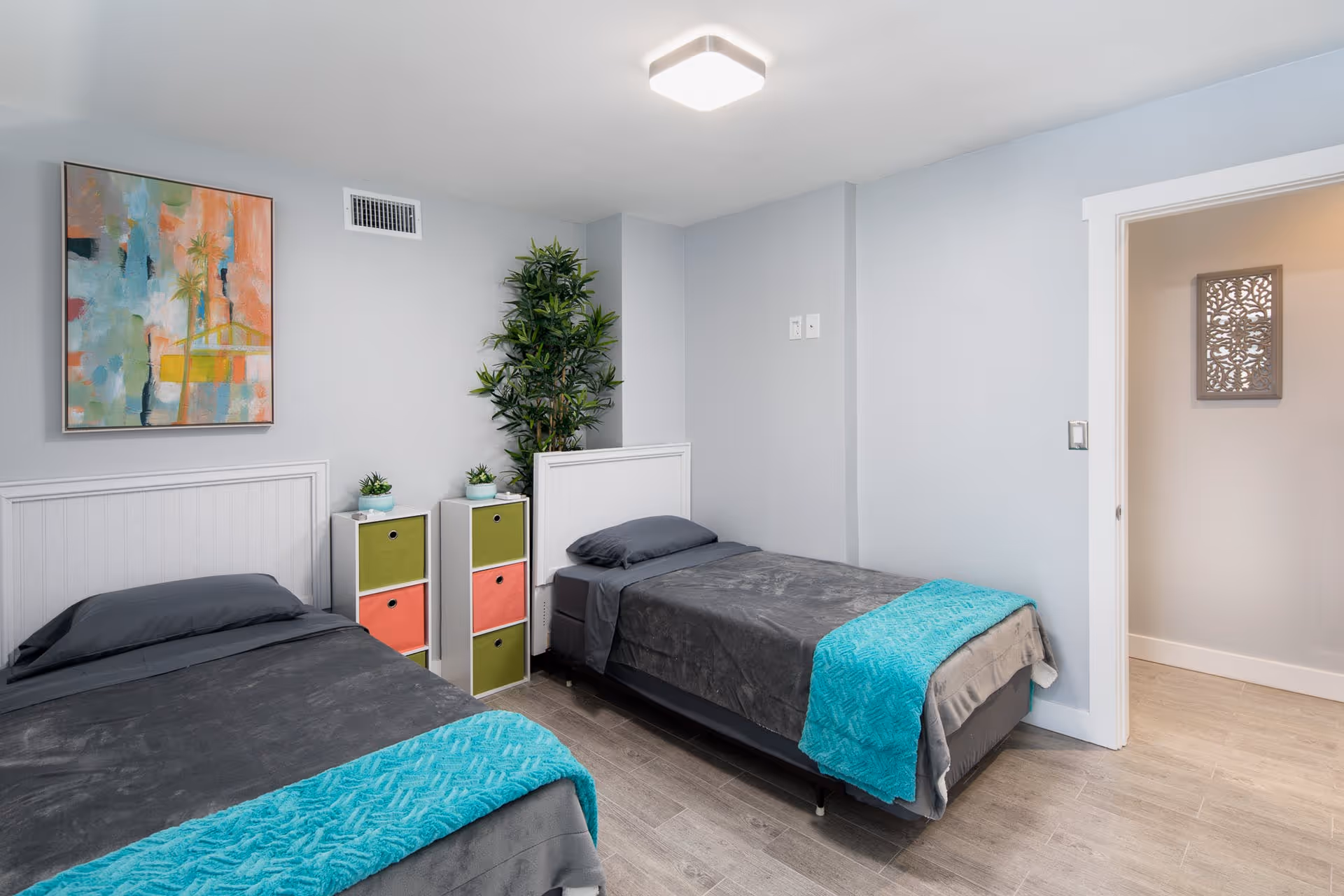 A clean and modern bedroom with two single beds, each covered with dark gray bedding and turquoise blankets at the foot. Between the beds is a small white storage unit with colorful drawers and small potted plants on top. A tall green plant is placed in the corner against a light gray wall. A colorful abstract painting hangs above one bed, and a doorway leads to another room with a decorative wall piece visible.