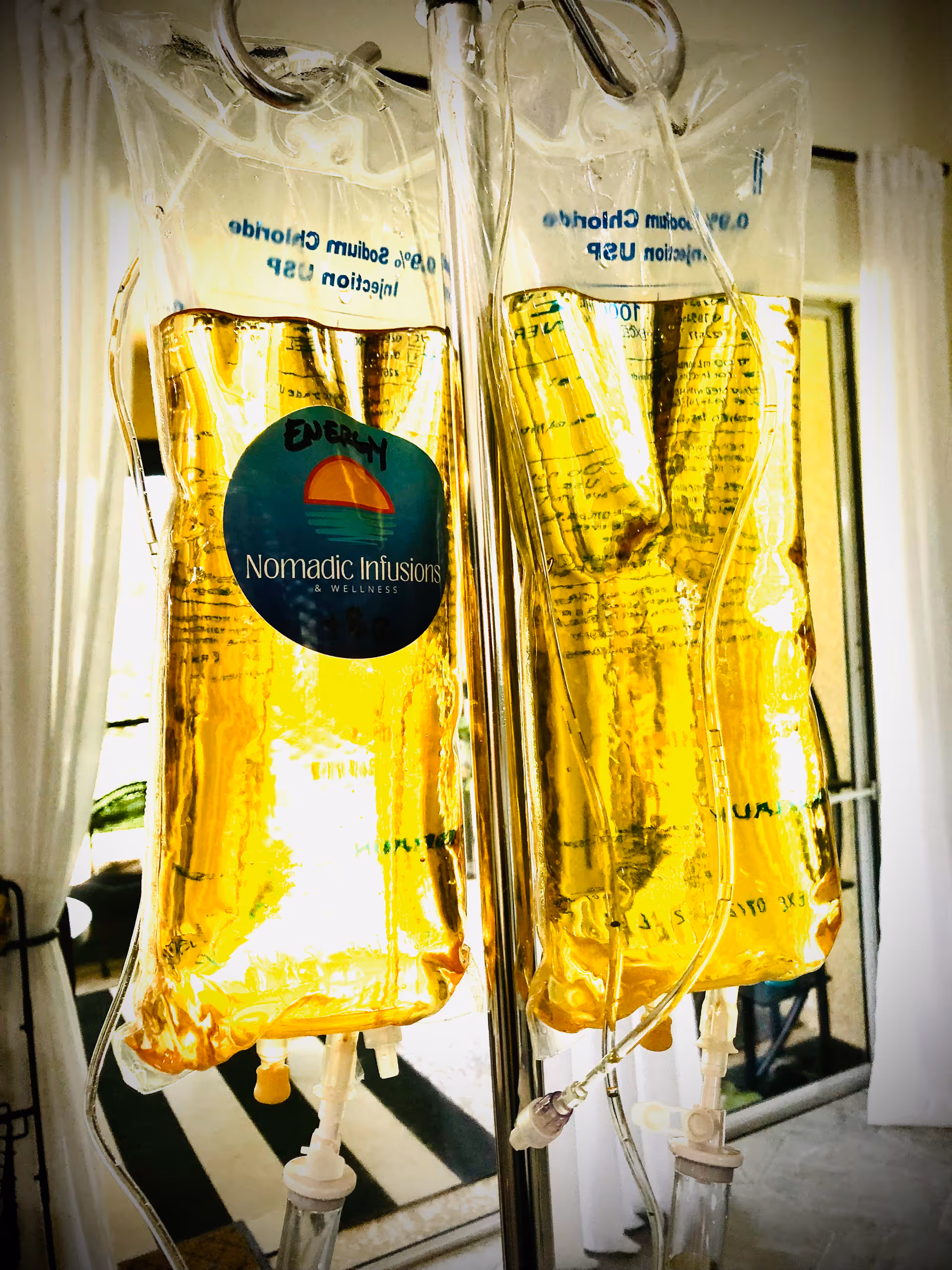 IV bags hang on a metal stand, labeled Nomadic Infusions & Wellness, in a medical interior.