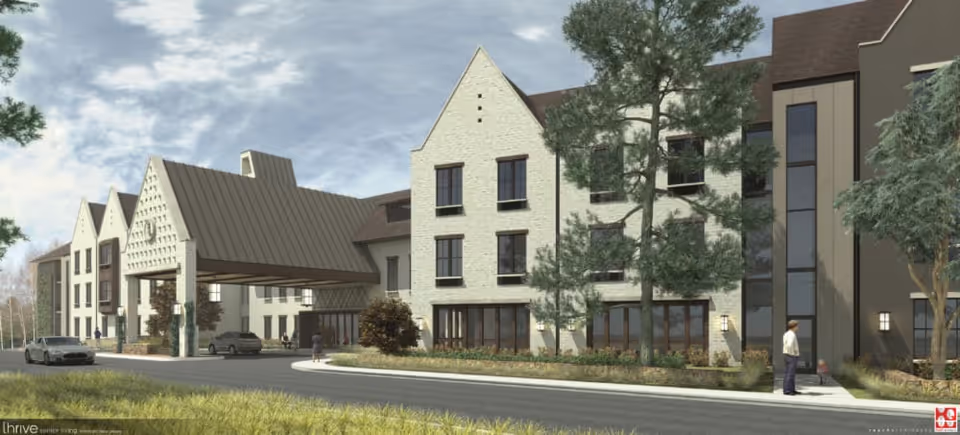 Front exterior of a multi-story senior living building with a covered entrance, driveway, landscaping, and parked cars.