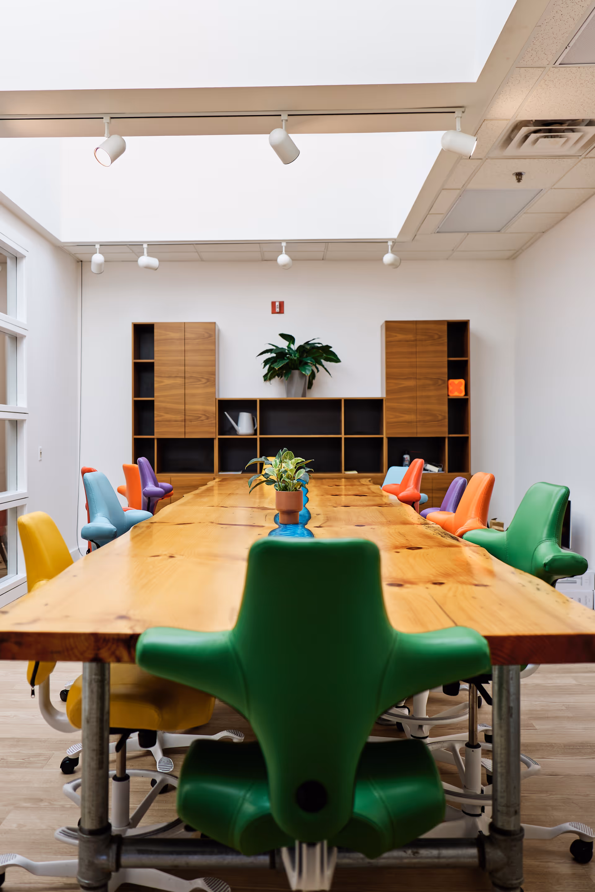 Long wooden conference-style table with colorful chairs in a bright, modern meeting room.