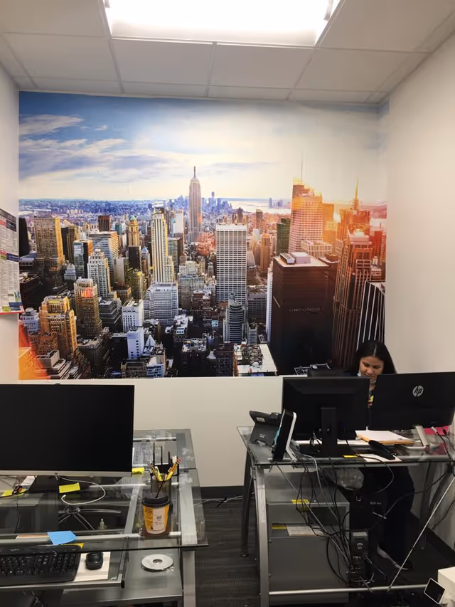 Office workspace with glass desks, computers, and a large cityscape mural on the wall.