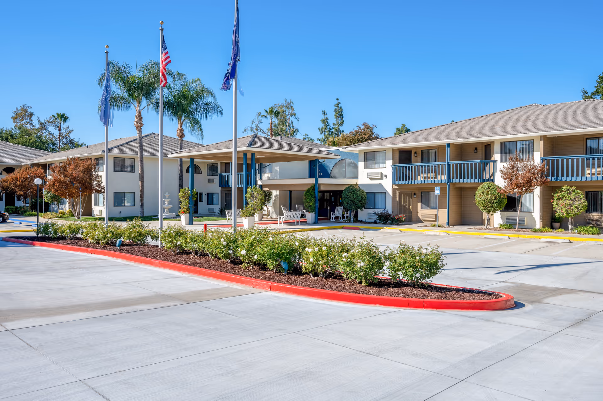 Exterior view of a senior living facility with two-story buildings, balconies, palm trees, and a covered entrance with seating. There are three flagpoles with flags in front of the building and a landscaped area with bushes and flowers along the driveway.