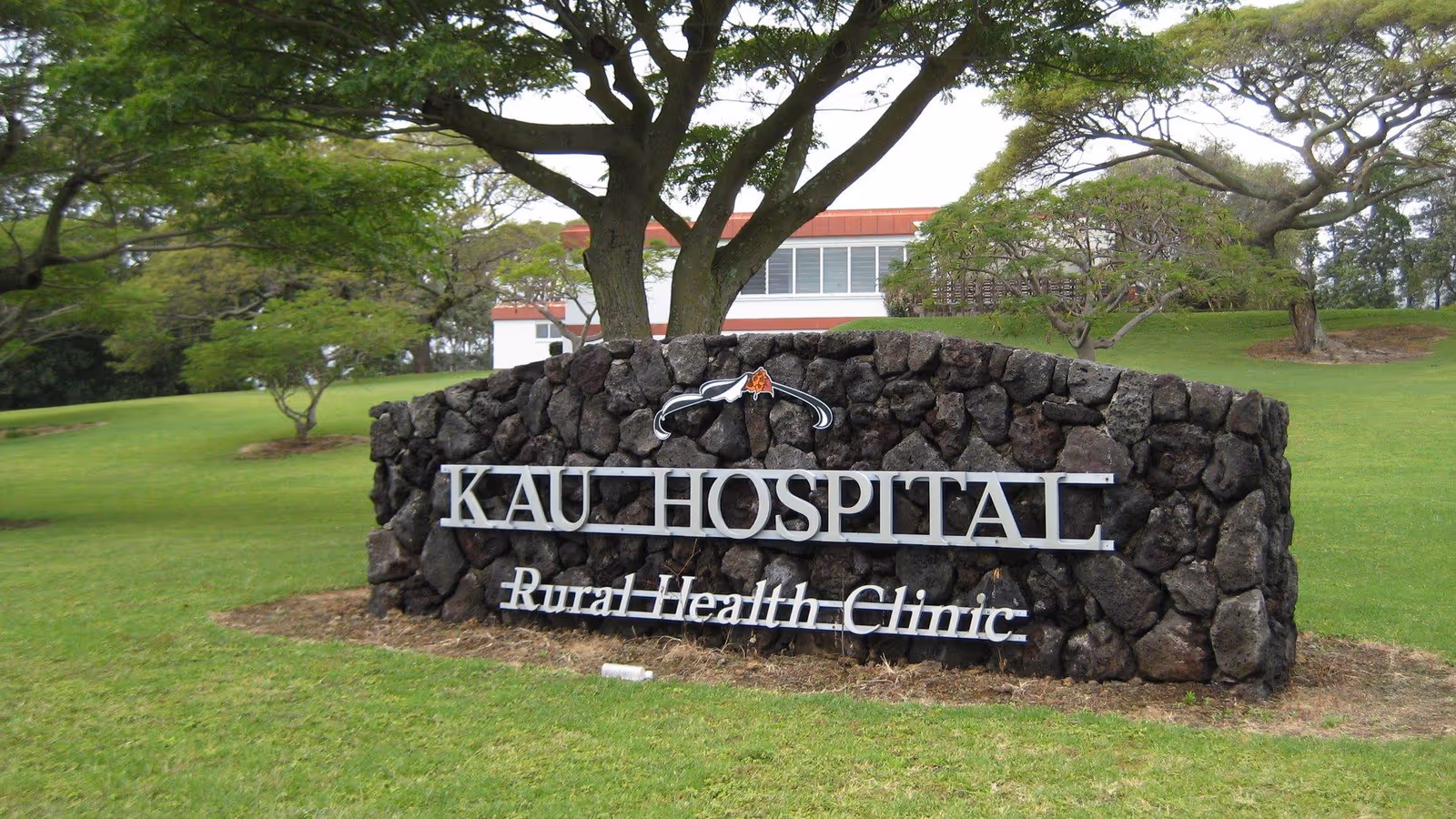 Stone sign reading Kaʻu Hospital Rural Health Clinic with green lawn and trees.
