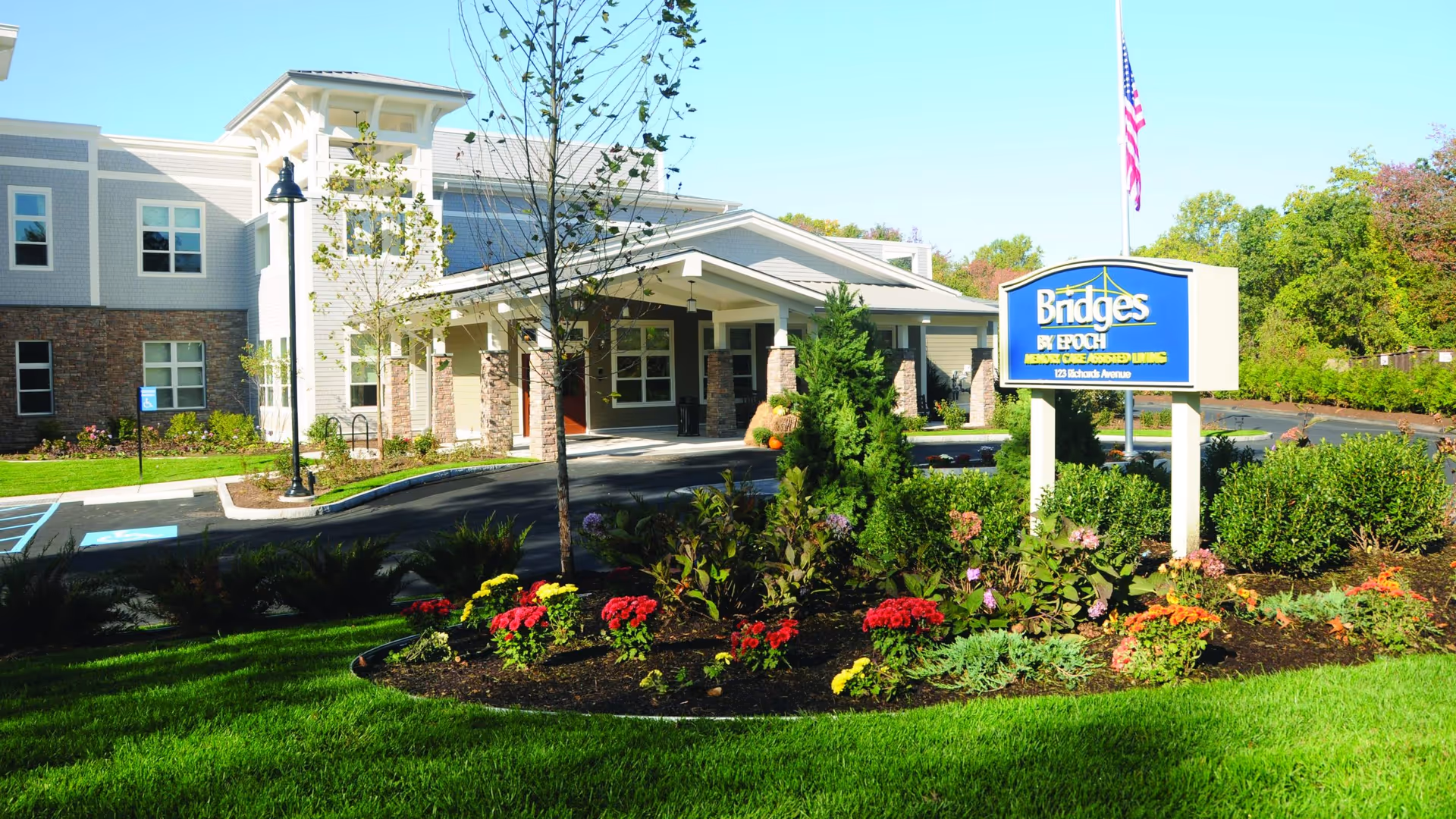 Exterior view of Bridges by EPOCH at Norwalk, a memory care assisted living facility, showing the building entrance with a covered driveway, landscaped garden with colorful flowers, and a sign with the facility name and address. An American flag is visible on a flagpole behind the sign.