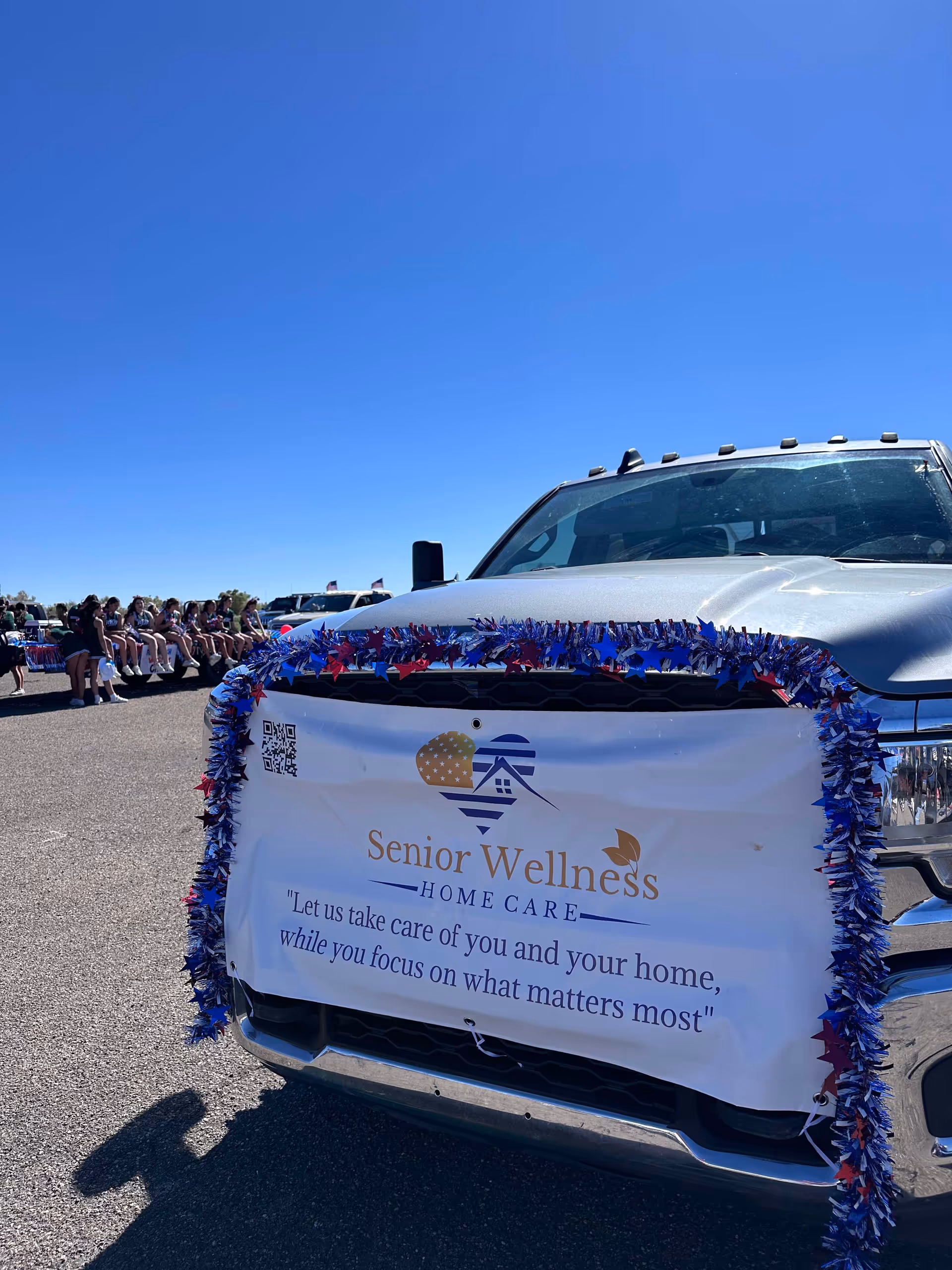 Silver pickup truck with Senior Wellness Home Care banner and blue garland at an outdoor event.