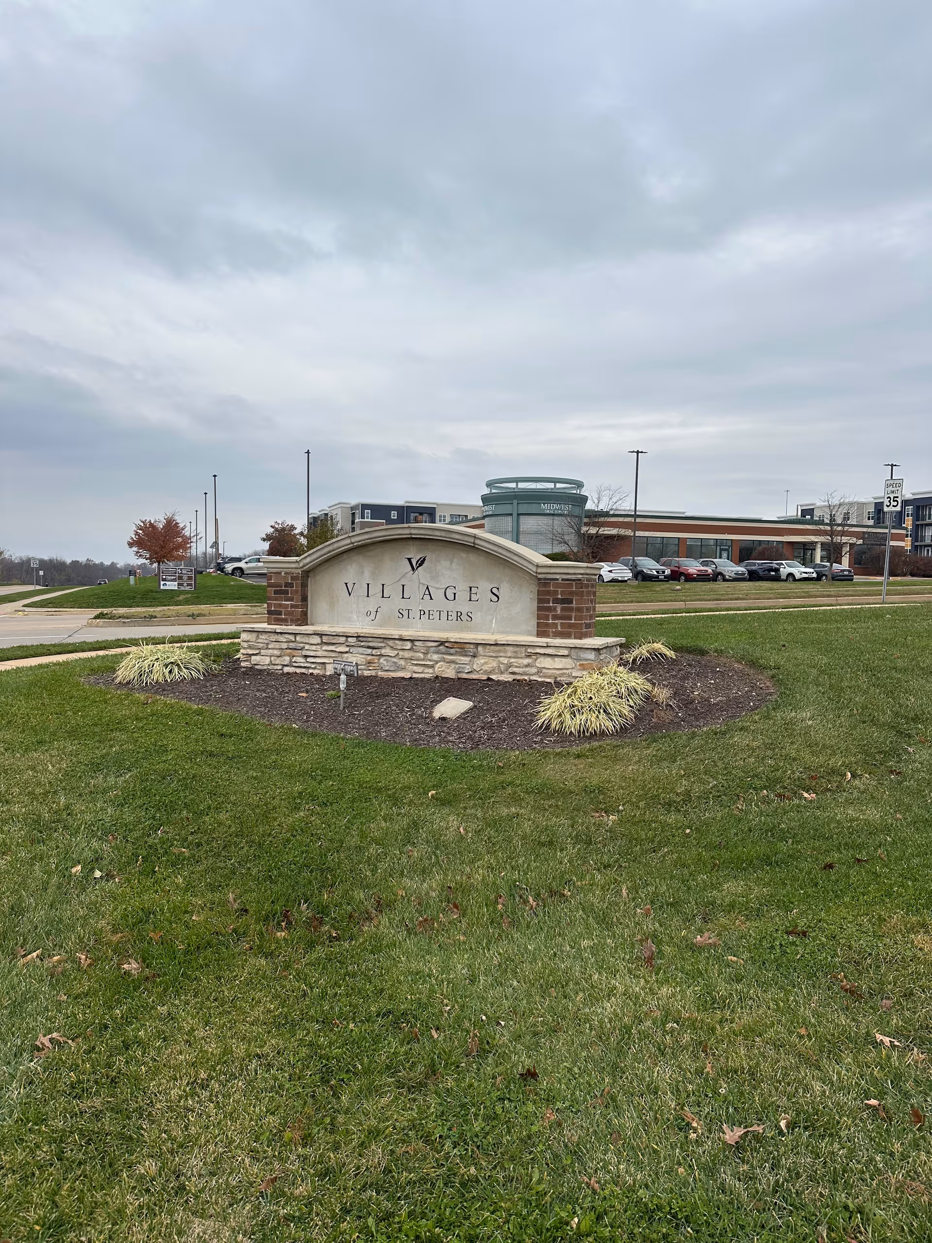 A stone and brick sign for Villages of St. Peters on a grassy area with a parking lot and buildings in the background under a cloudy sky.