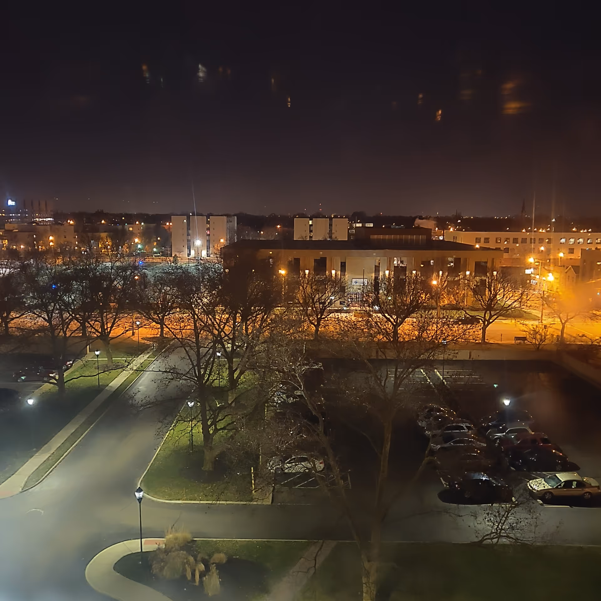 Nighttime exterior view of a facility with parking, bare trees, and streetlights.