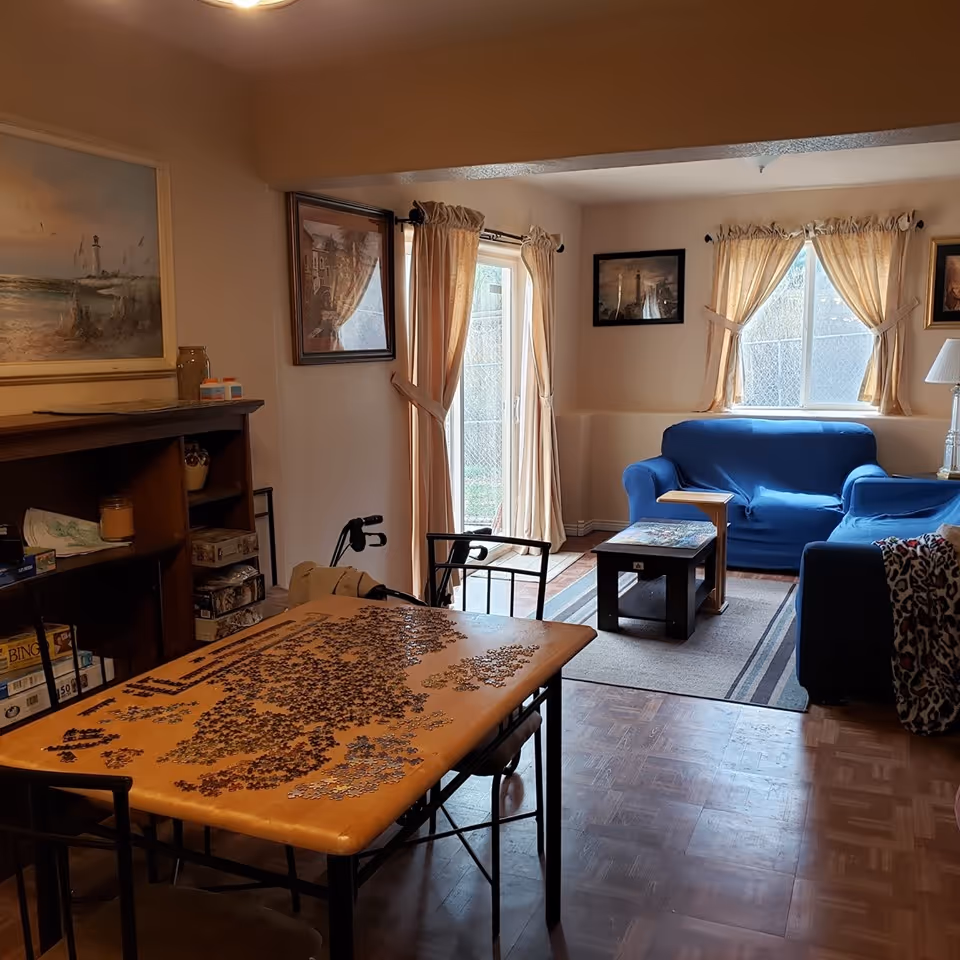 Cozy living room with blue sofa, windows with curtains, wooden puzzle table, and walker nearby.