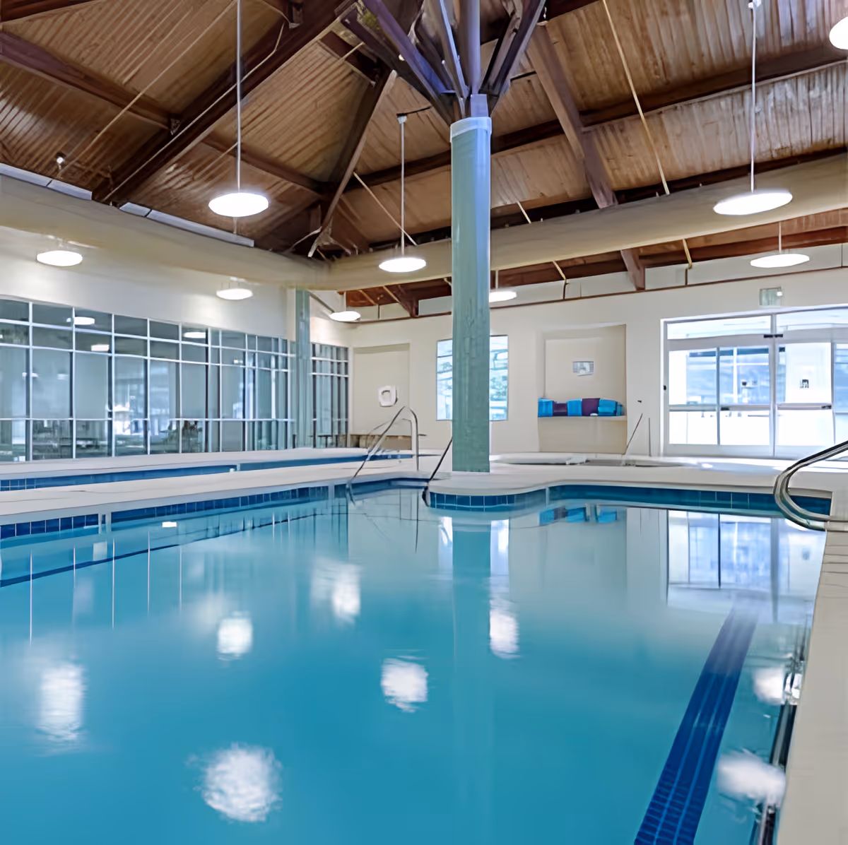 Indoor swimming pool with clear blue water, surrounded by white tiled flooring and a high wooden ceiling with exposed beams and hanging round lights. Large windows and glass doors allow natural light to enter the space.