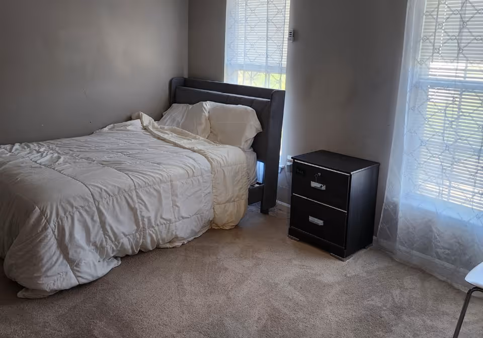 Bedroom with a white comforter, dark nightstand, and windows covered by sheer curtains
