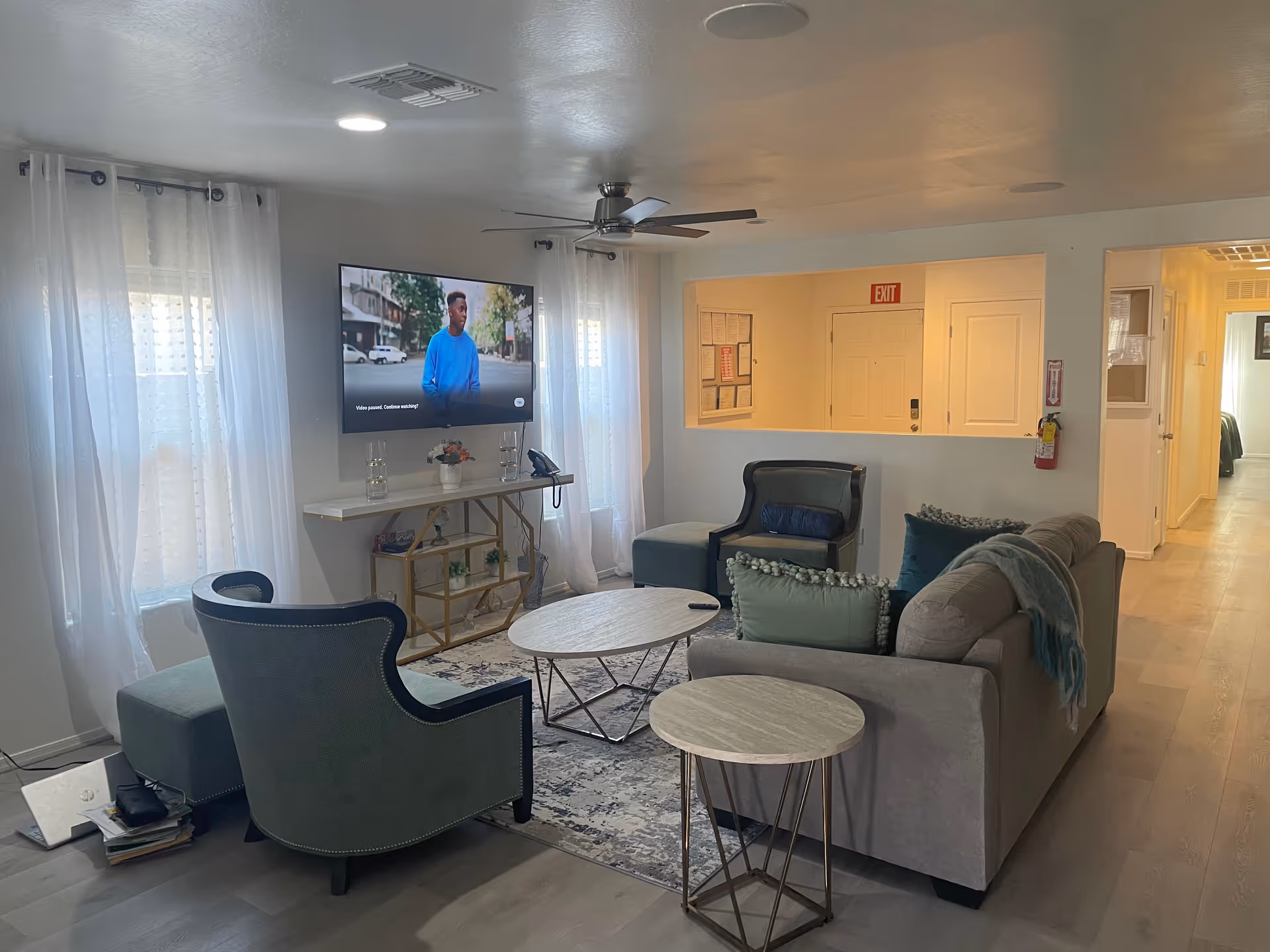 Bright communal living room in a senior facility with sofa, seating, TV, and sheer curtains.