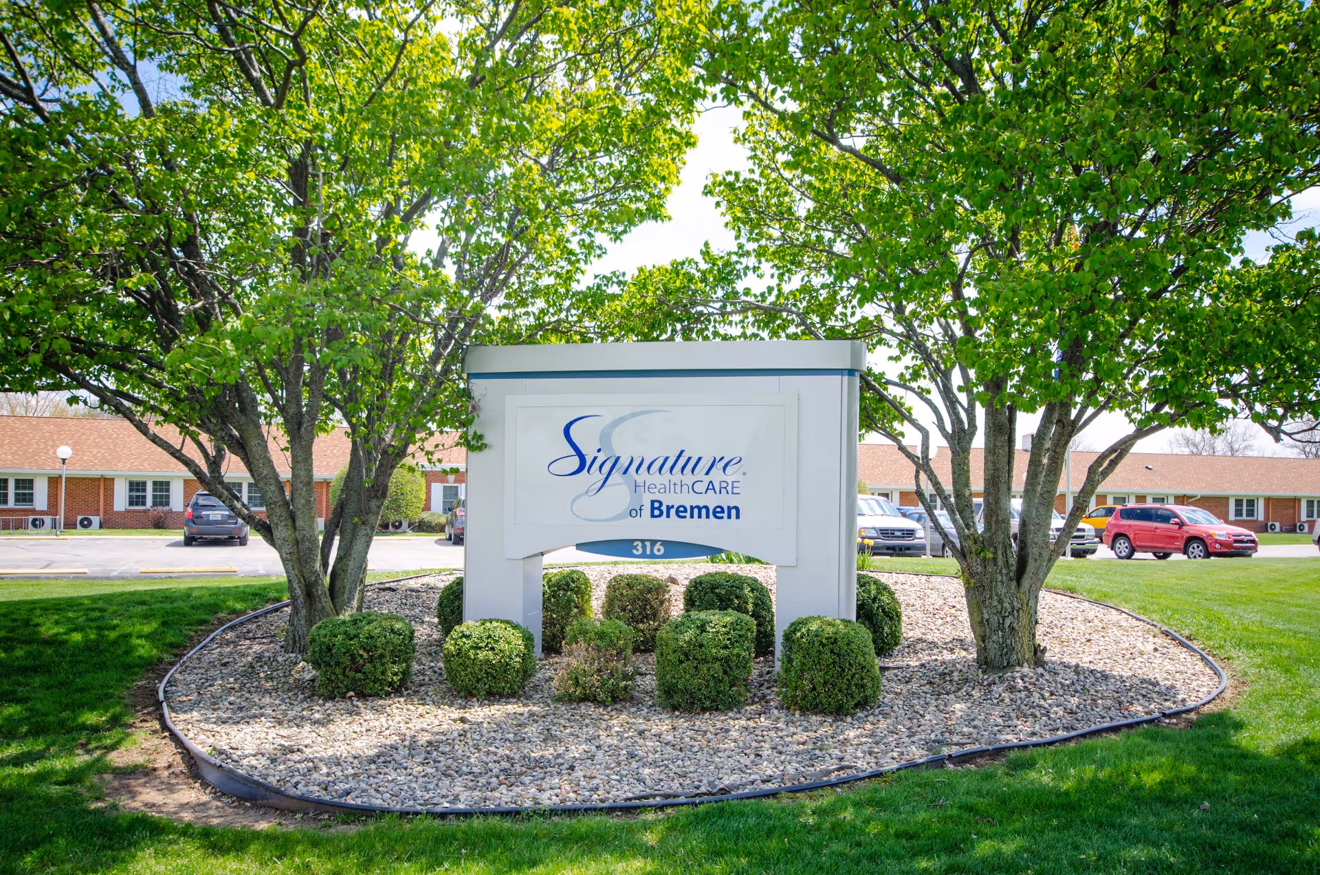 Sign for 'Signature HealthCARE of Bremen' in a landscaped island with trees and the facility building and parked cars behind it.