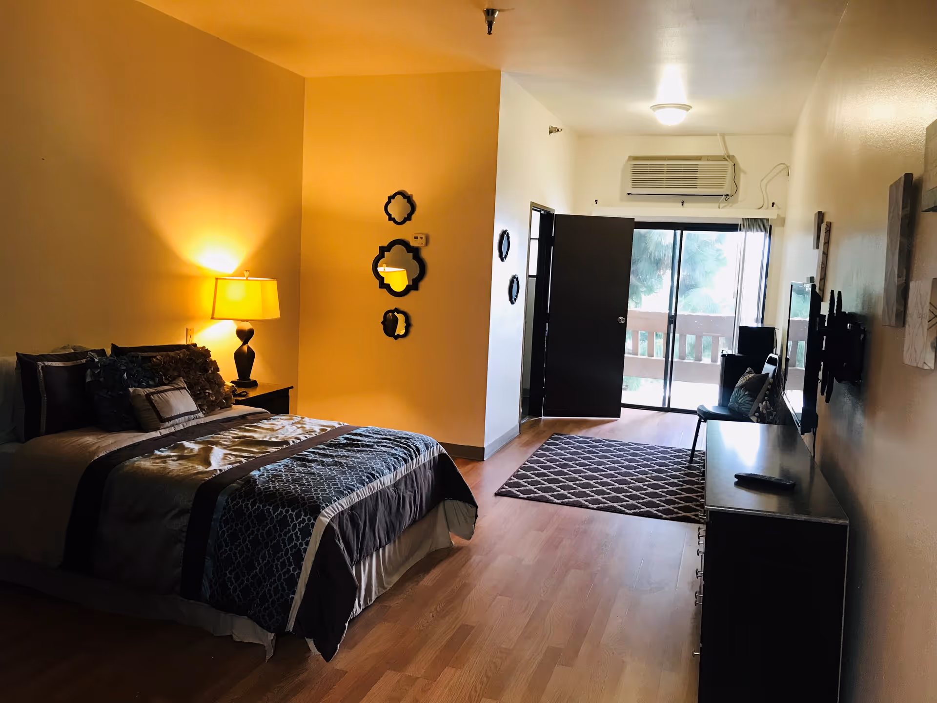 Furnished bedroom in a retirement center with bed, nightstand, dresser, and sliding balcony door.