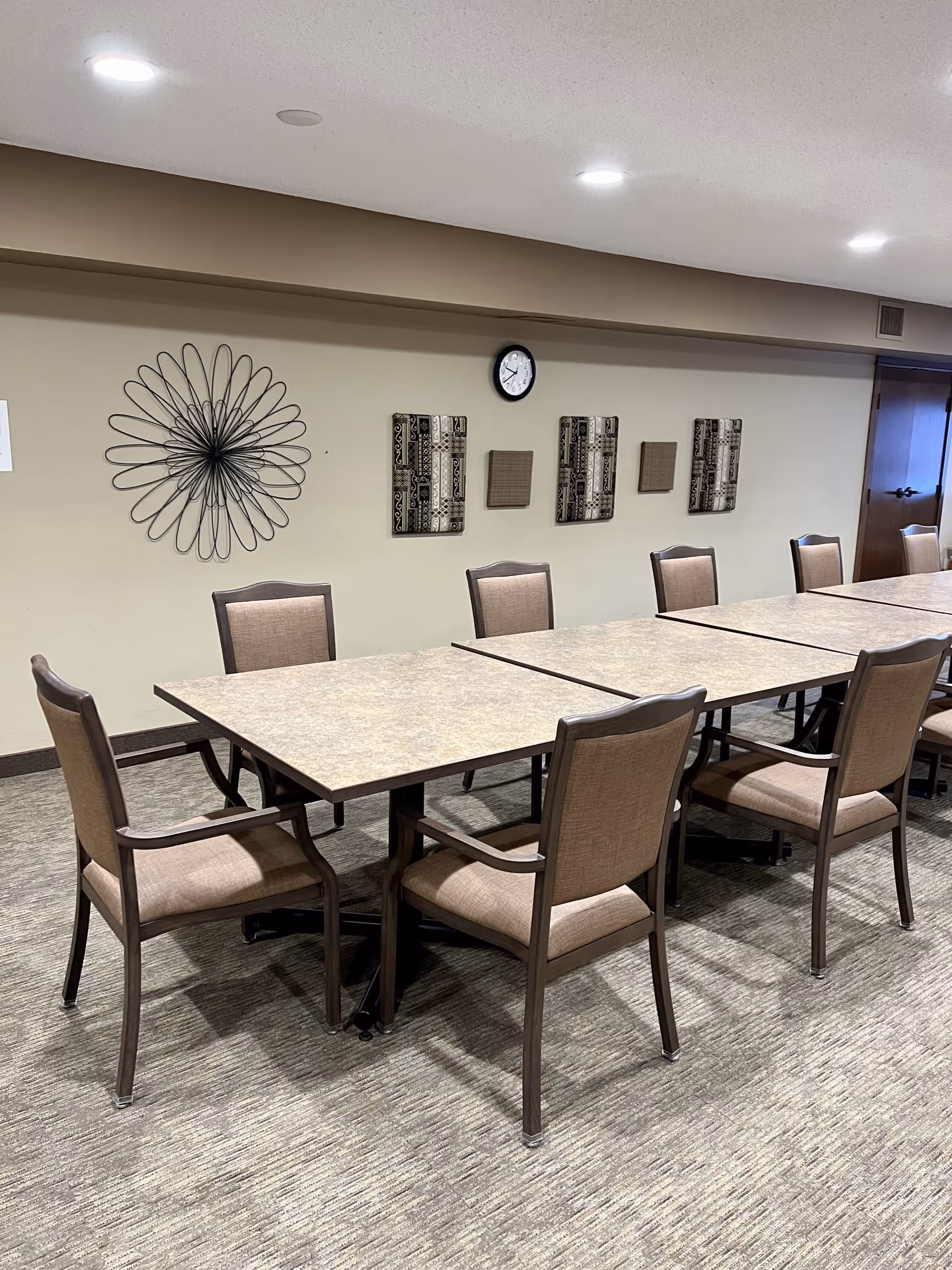 Long rectangular tables with padded chairs arranged in a communal dining/activity room with wall decor and a clock.