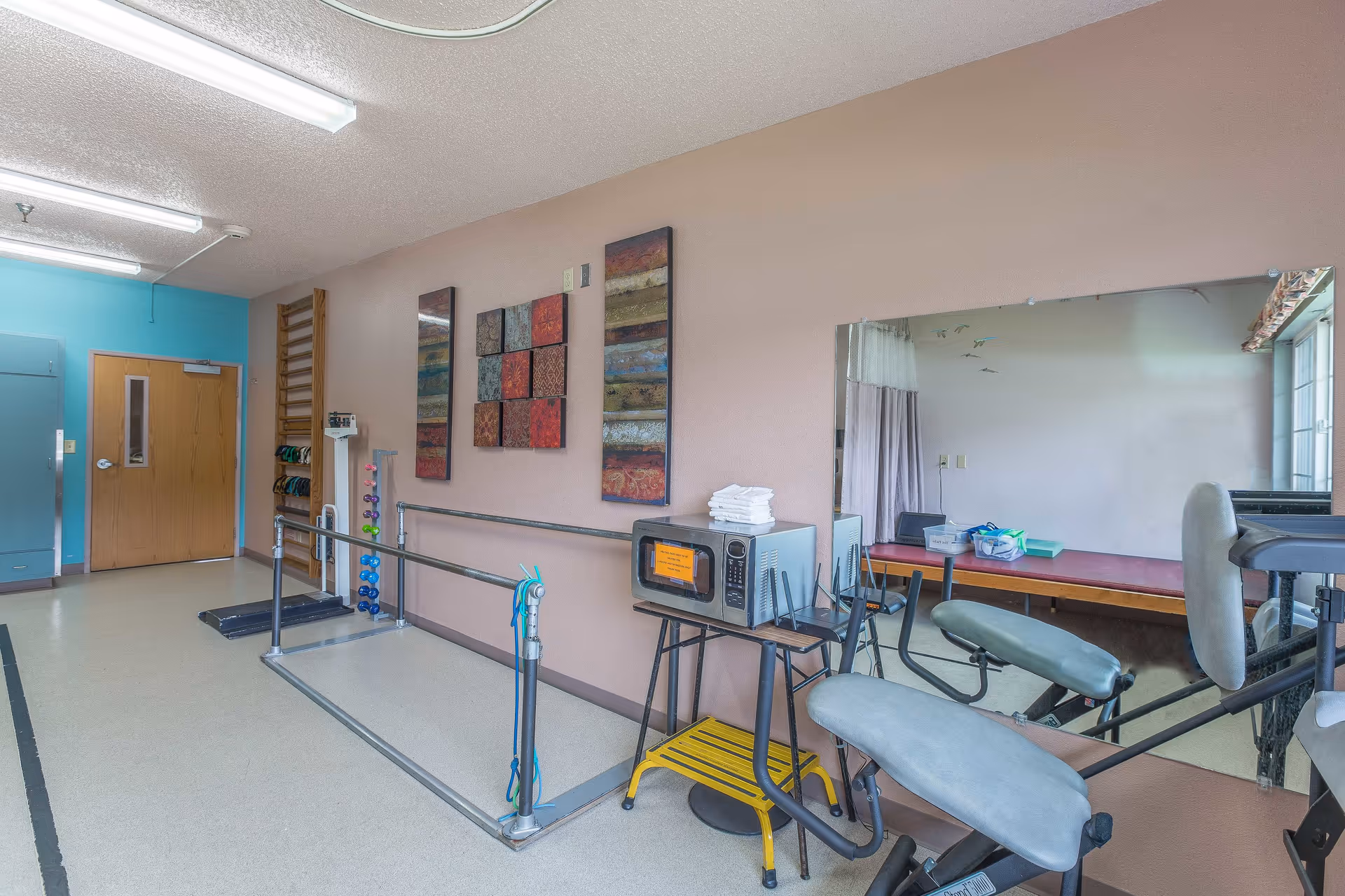 Rehab gym featuring parallel bars, exercise equipment, and a microwave on a cart.