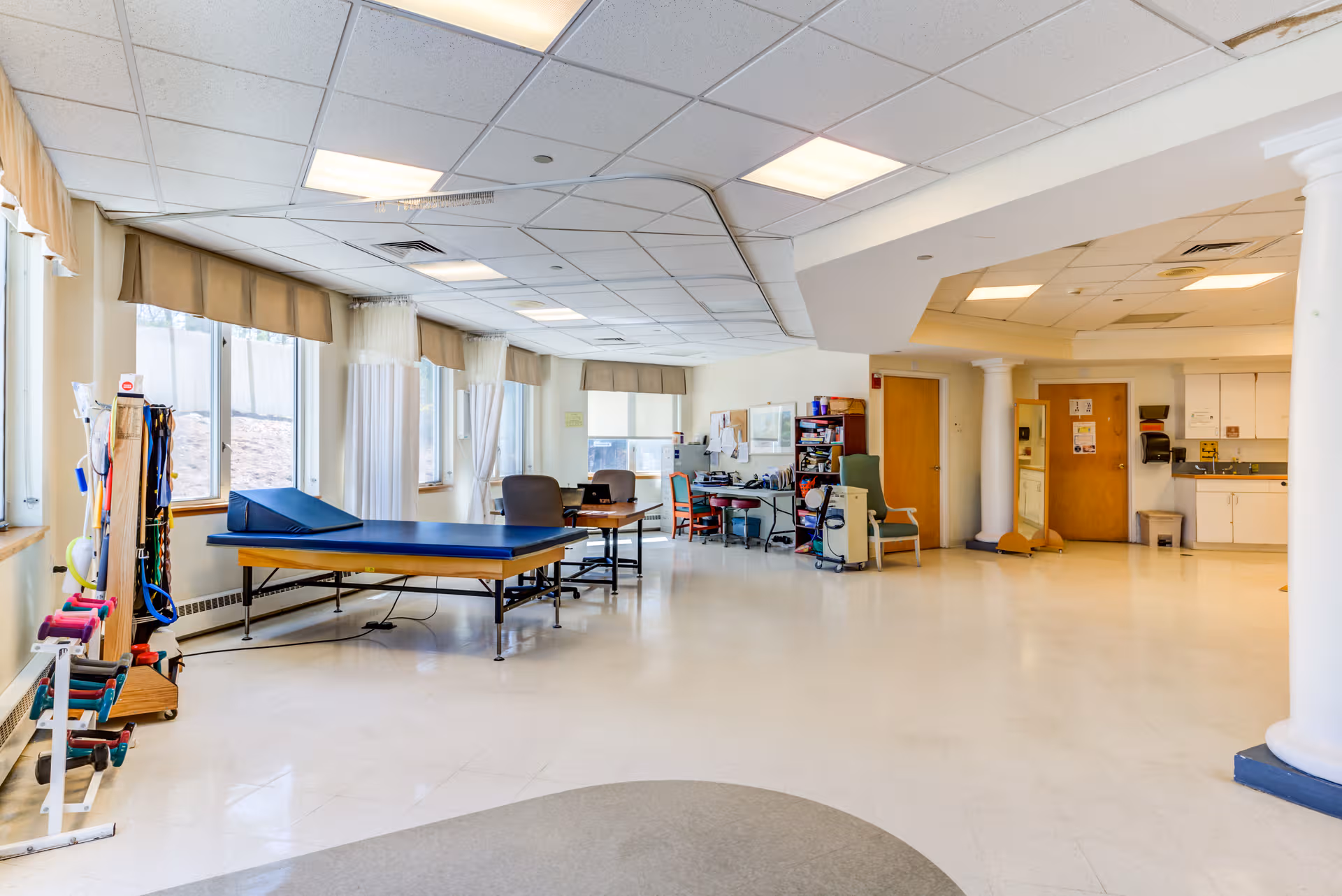 Bright rehabilitation room with therapy table, chairs, equipment, and kitchenette in back.