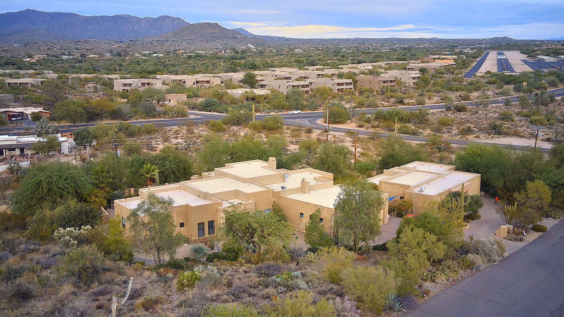 Aerial view of a beige stucco assisted living complex with flat roofs surrounded by desert landscaping and distant mountains.