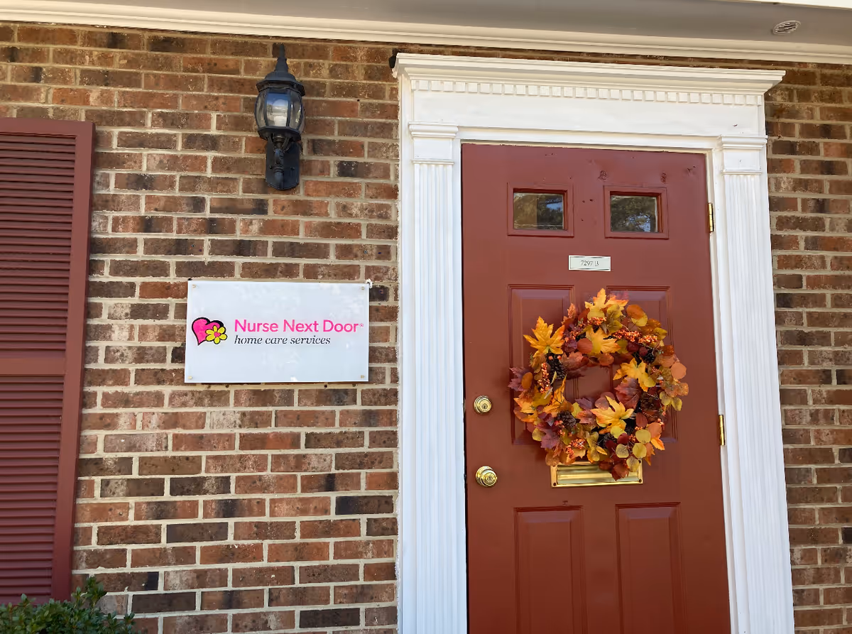 Brick building exterior with a red front door, wreath, lantern, and Nurse Next Door sign.