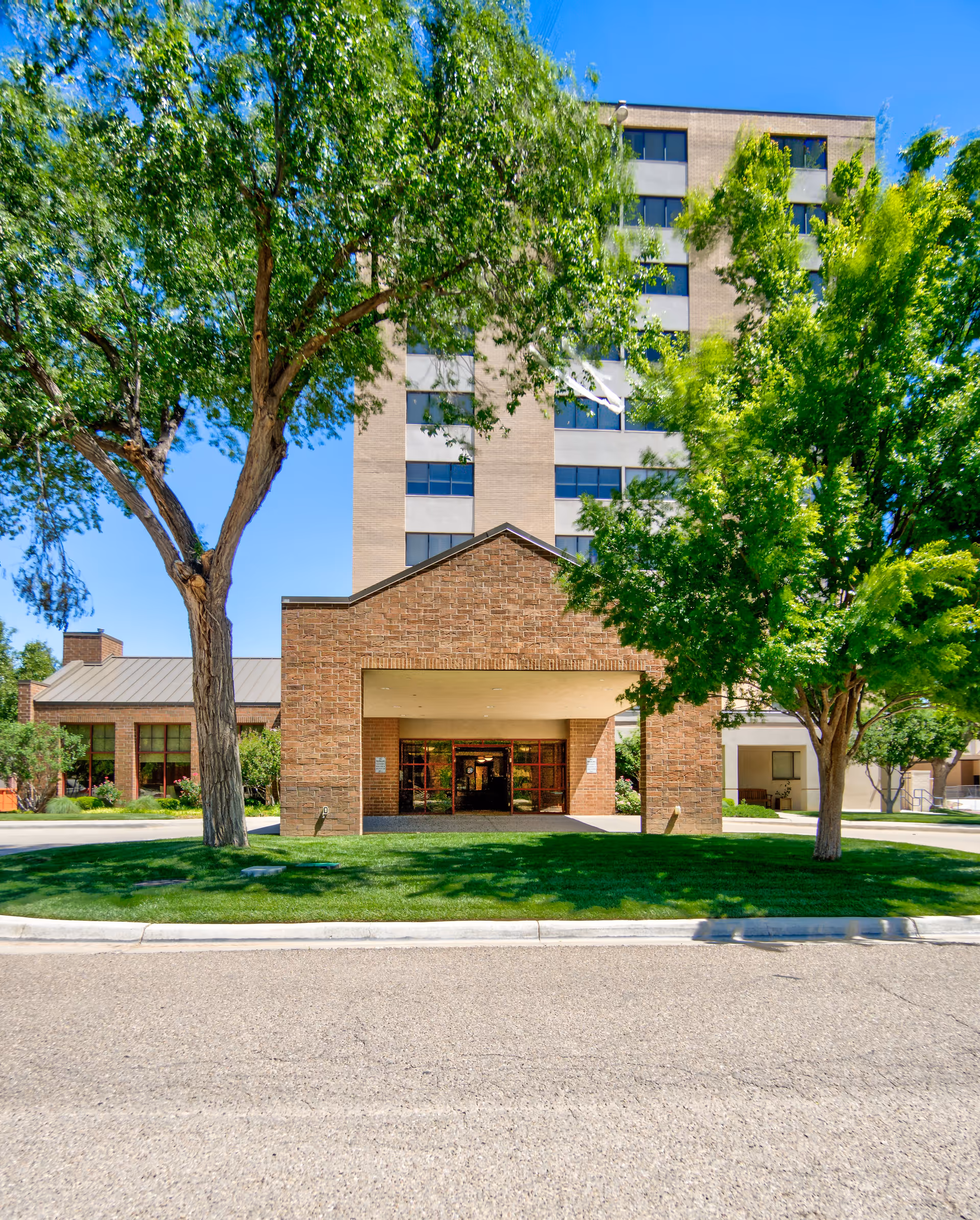 Brick-faced senior living facility entrance beneath green trees on a sunny day