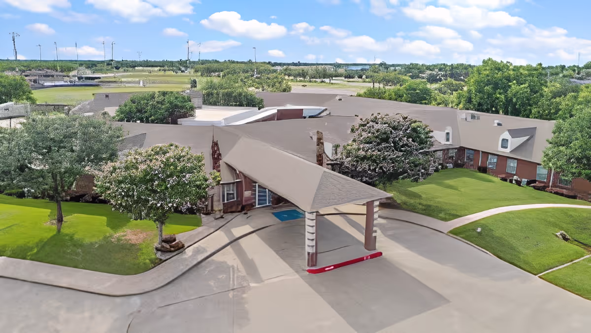 Aerial view of Morada Abilene senior living facility showing a single-story building with a covered entrance, surrounded by green lawns, trees, and a clear blue sky with some clouds.