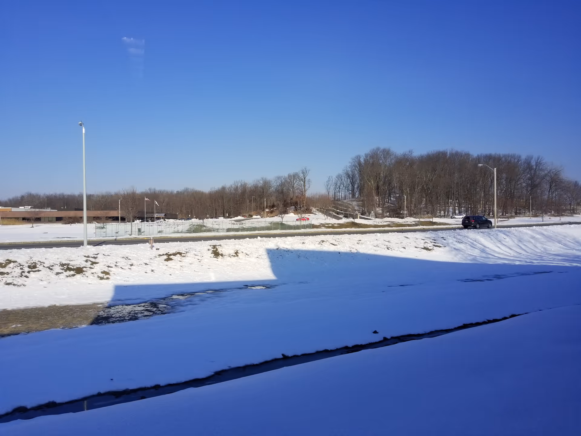 Snow-covered parking area and road with leafless trees under a blue sky.