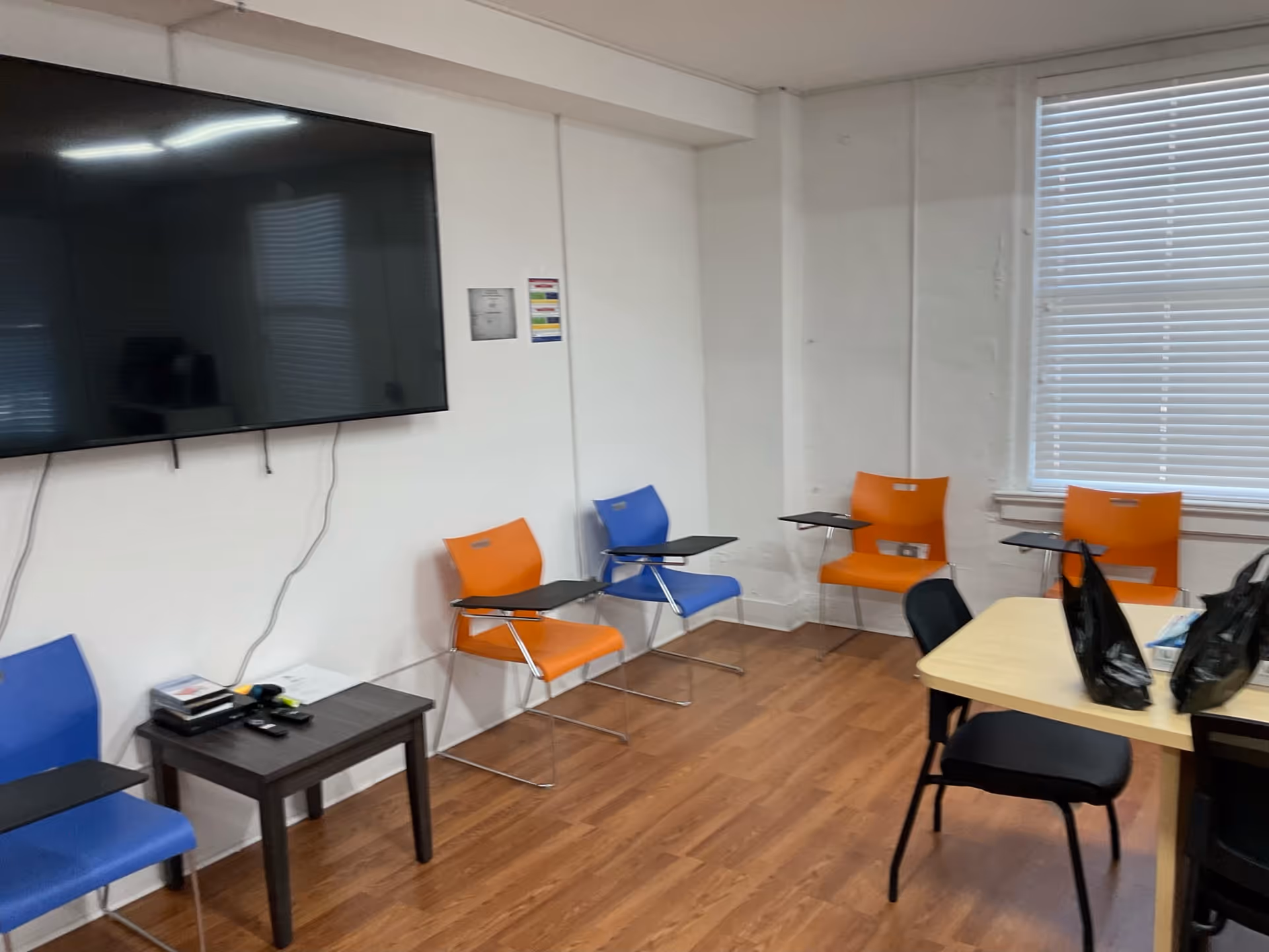 A small interior room with orange and blue chairs, desks, and a wall-mounted TV.