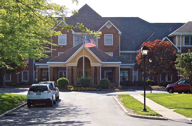 Front exterior view of a brick senior living facility with a peaked roof and an American flag flying on a flagpole in front. There are cars parked along the driveway and well-maintained landscaping with trees and bushes around the building.