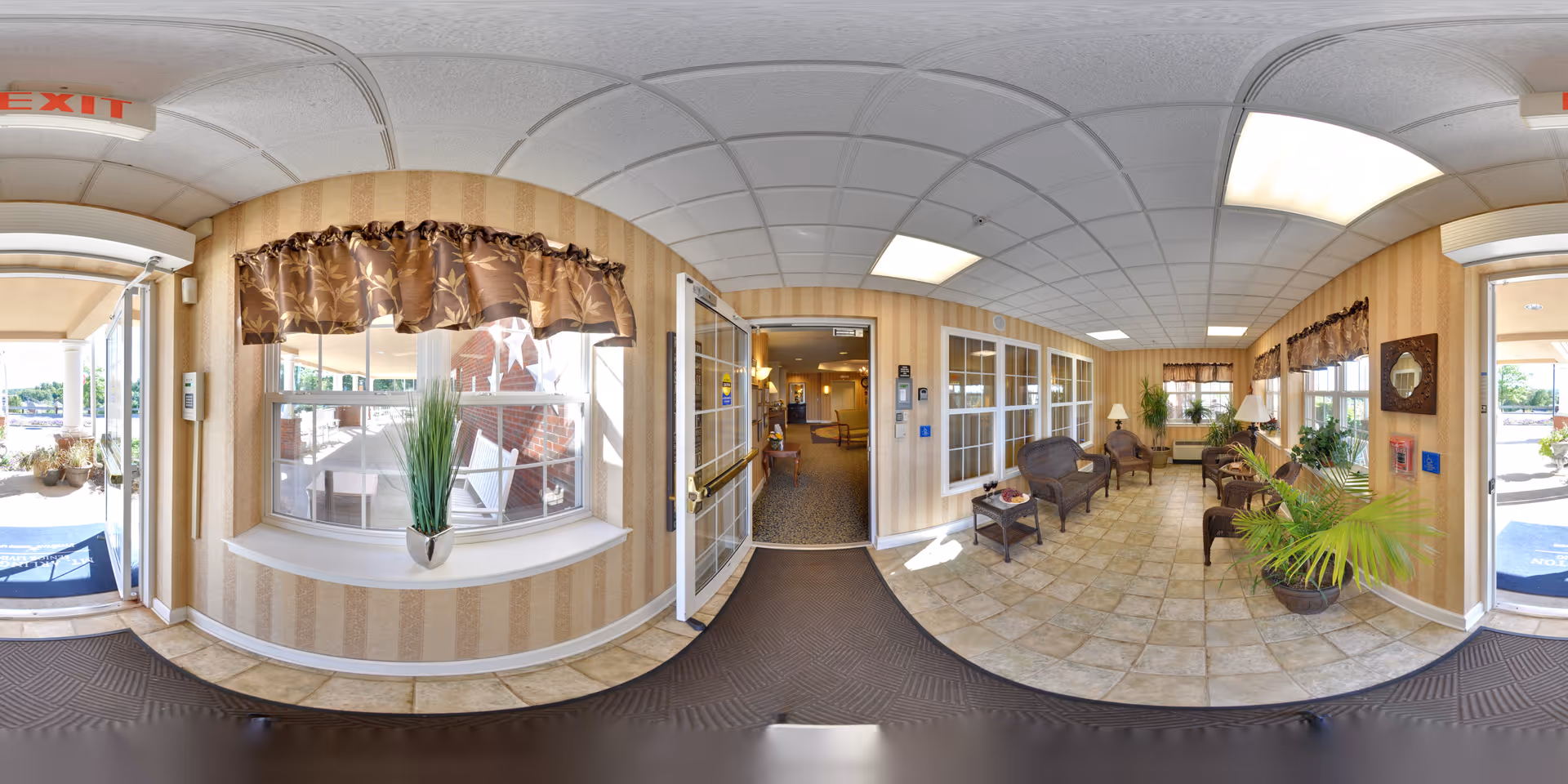 Entrance area of Mt. Arlington Senior Living facility showing a tiled floor hallway with several wicker chairs and small tables along the walls. There are large windows with brown patterned valances, potted plants, and lamps providing a welcoming atmosphere. Double glass doors lead into an interior space, and an exit sign is visible above the door to the outside.