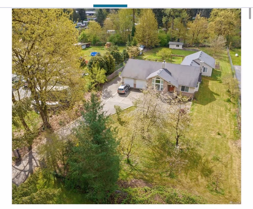 Aerial view of a single-story residential building with a gray roof surrounded by green trees and grass. There is a driveway with a parked car in front of the house, and a fenced backyard with additional structures and greenery.