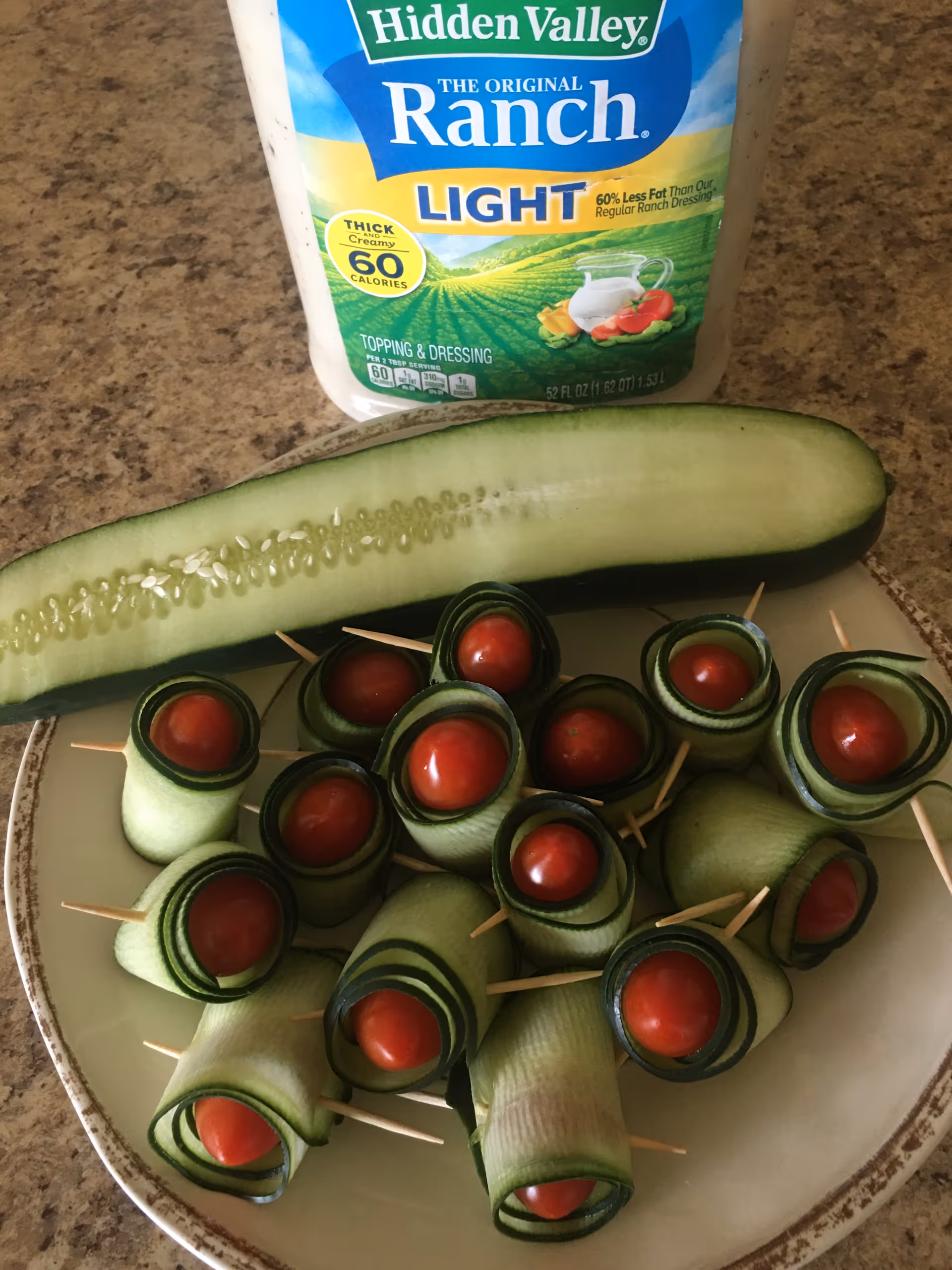 Cucumber canapés with cherry tomatoes on toothpicks arranged on a plate, ranch dressing bottle in background.