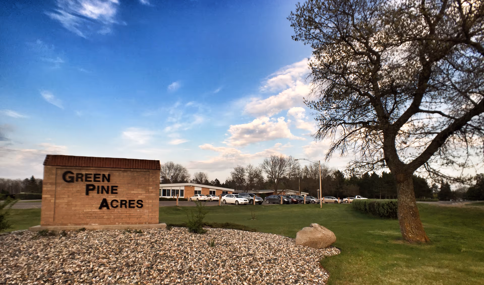Exterior view of Green Pine Acres facility with sign, parking lot, and a tree against sky.