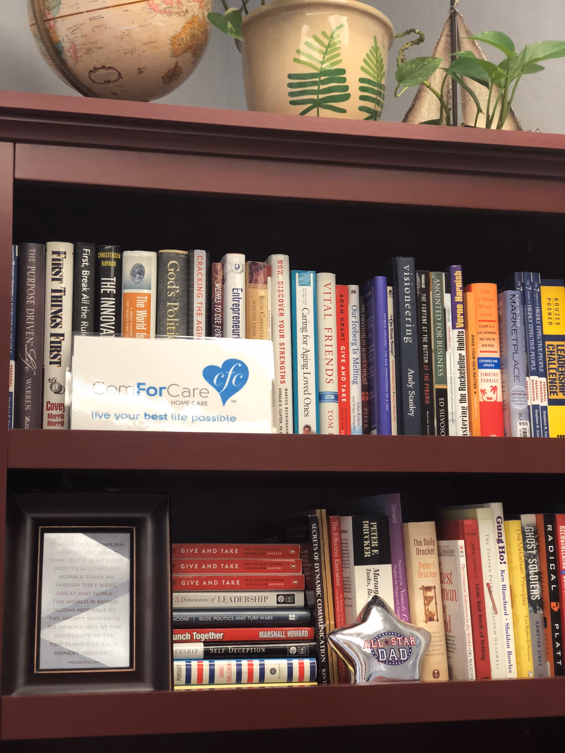 Interior bookshelf with many books, a ComForCare sign, plants, and a framed item.