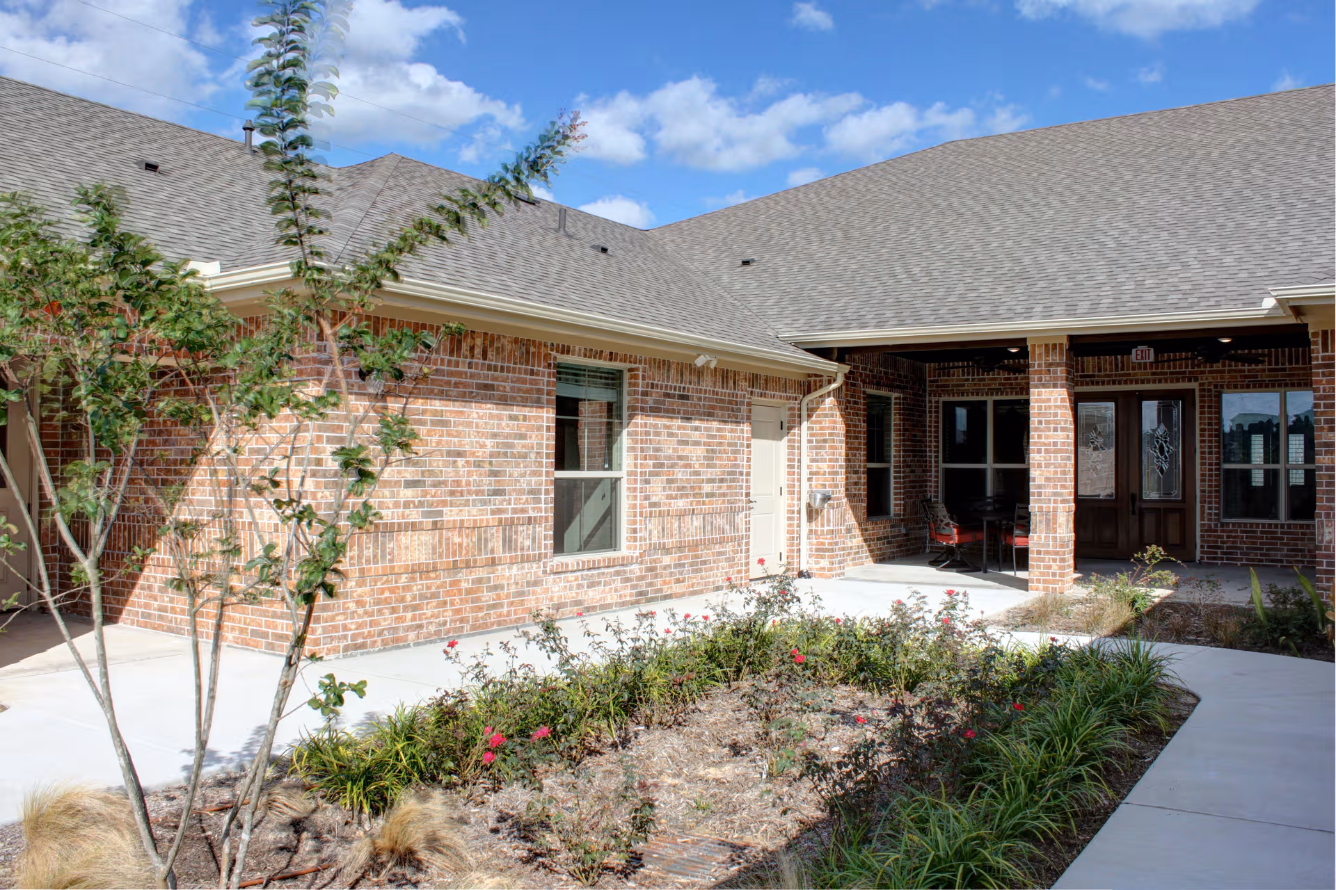 Brick senior living facility exterior with a covered entrance, landscaped walkway, and blue sky.