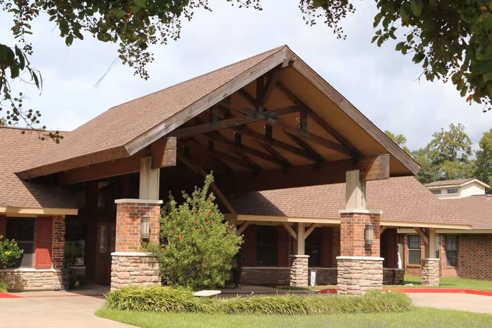 Exterior view of Timberwood Nursing & Rehabilitation Center showing a covered entrance with wooden beams, brick and stone pillars, and a well-maintained lawn with shrubs and trees.