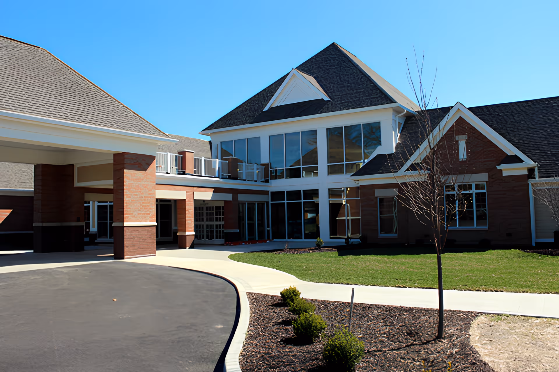 Entrance and front exterior of a two-story senior living building with a covered drive-through porte-cochere, large windows, and landscaped grounds.