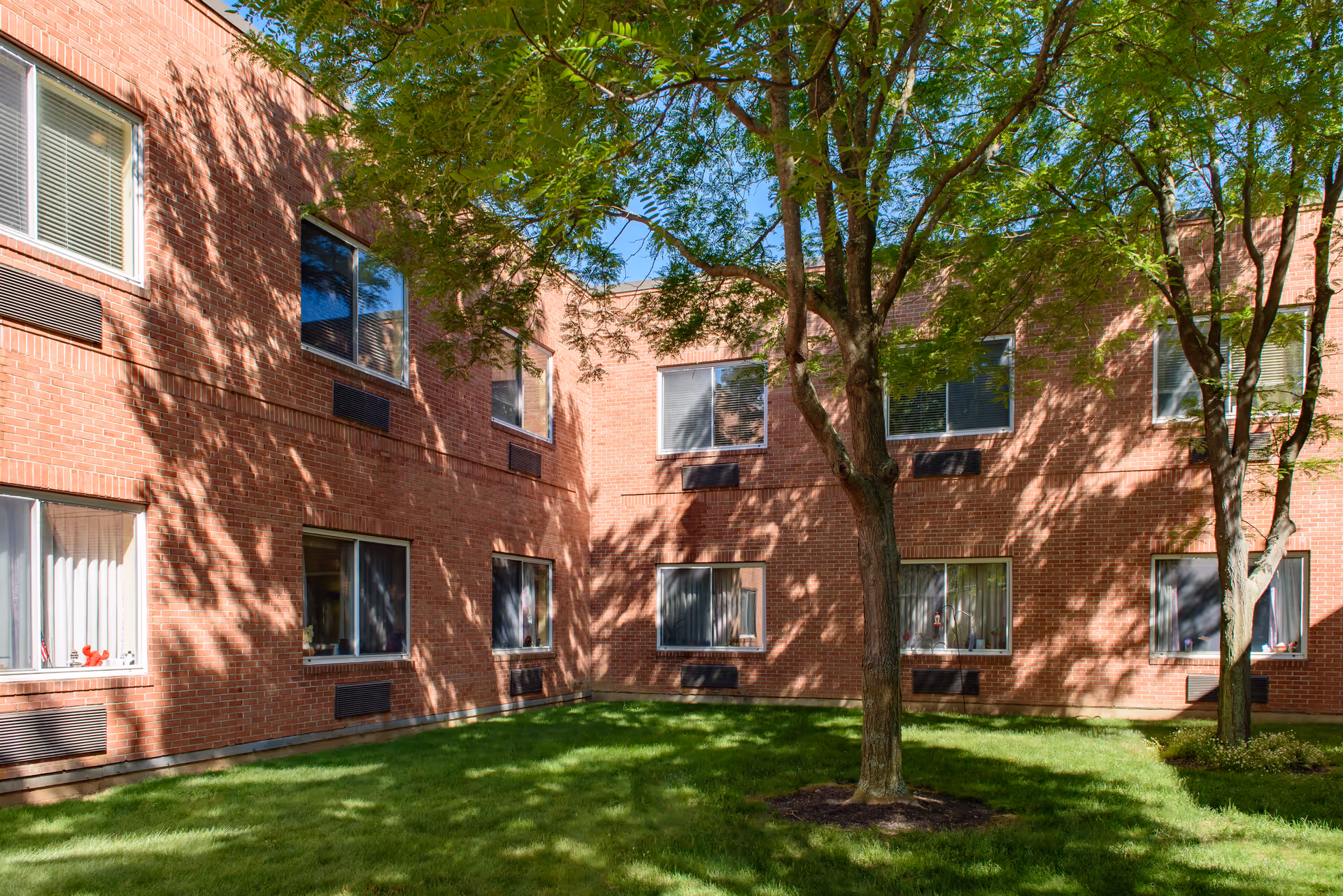Brick two-story building courtyard with windows, a grassy lawn and trees casting shadows.