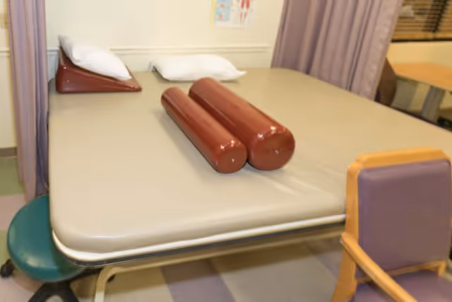 A therapy or examination table with two cylindrical brown bolsters and two white pillows placed on it. The table is in a room with a green stool, a wooden chair with purple cushion, and a curtain divider. There is a medical poster on the wall in the background.