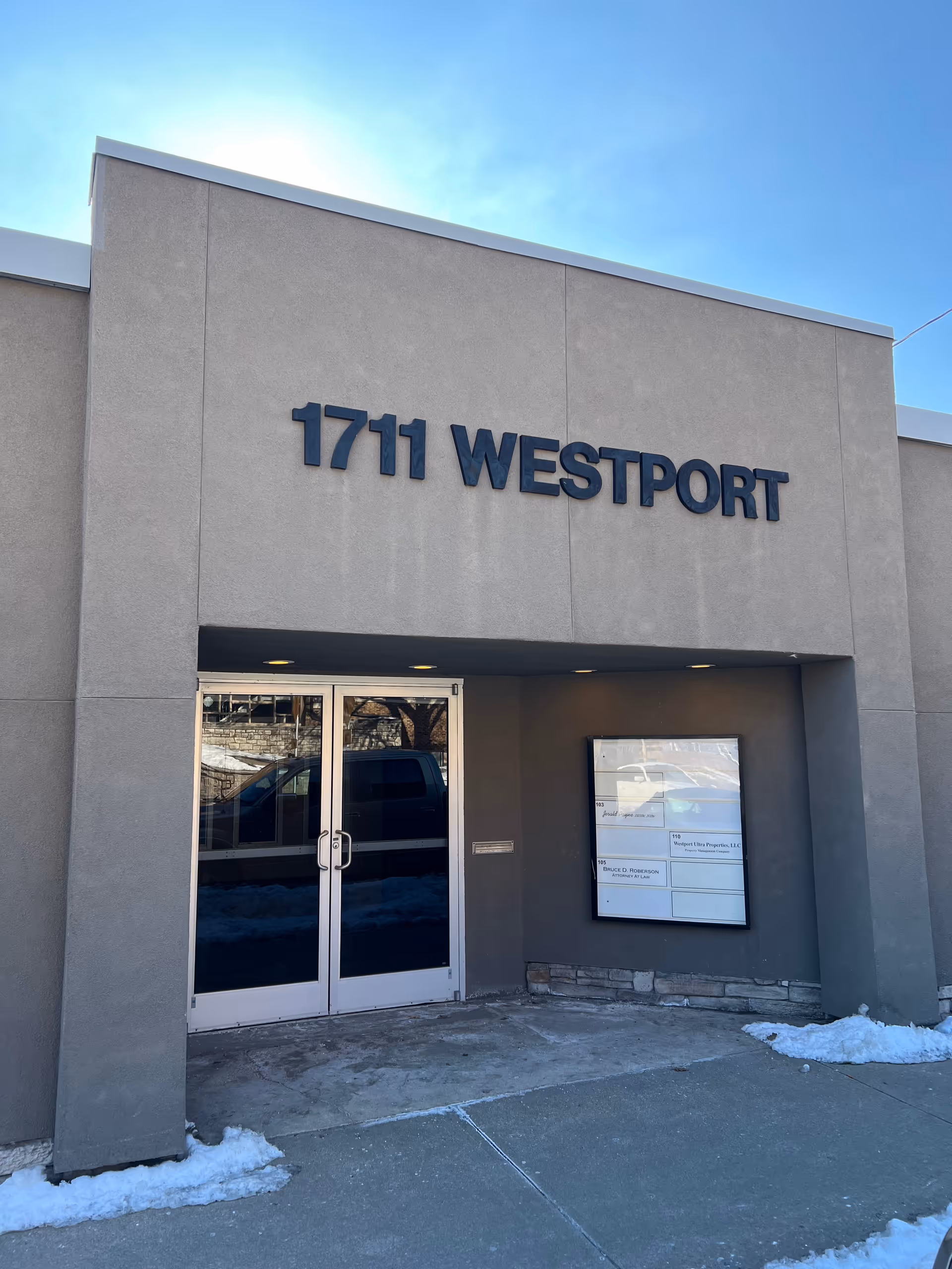 Grey commercial building with a large 1711 WESTPORT sign and glass double doors, snow on ground.