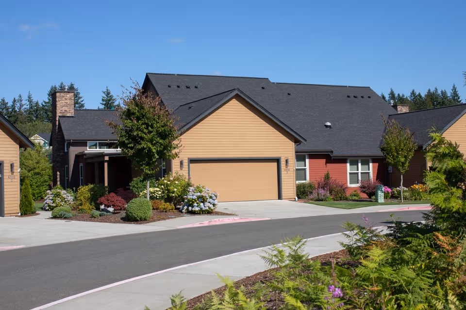 Front view of single-story attached homes with garages, landscaped gardens, and a curved road under a clear blue sky.