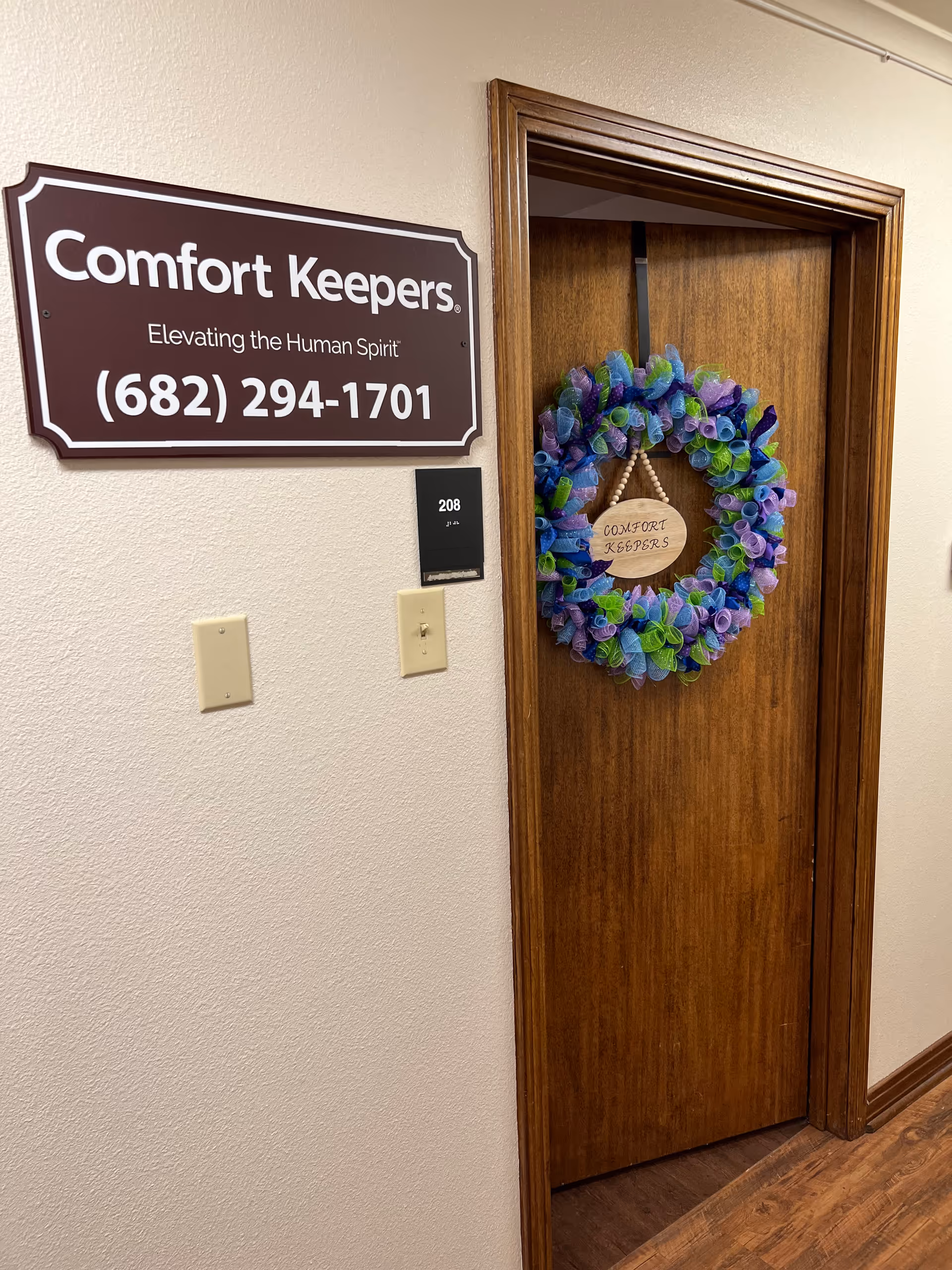 A hallway wall features a Comfort Keepers sign and a door with a colorful wreath.