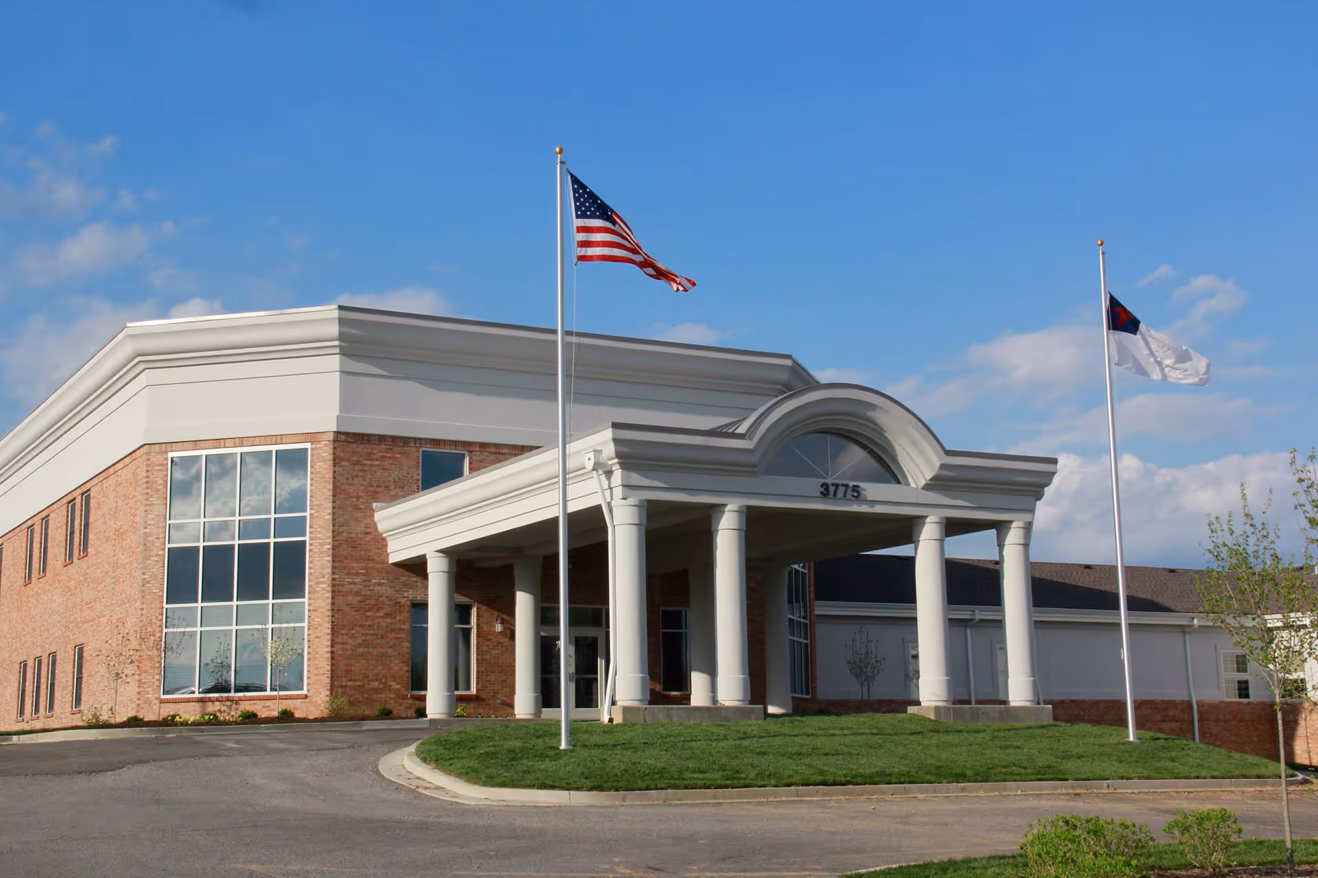 Entrance of a brick building with white columns and two flagpoles flying flags under a blue sky.