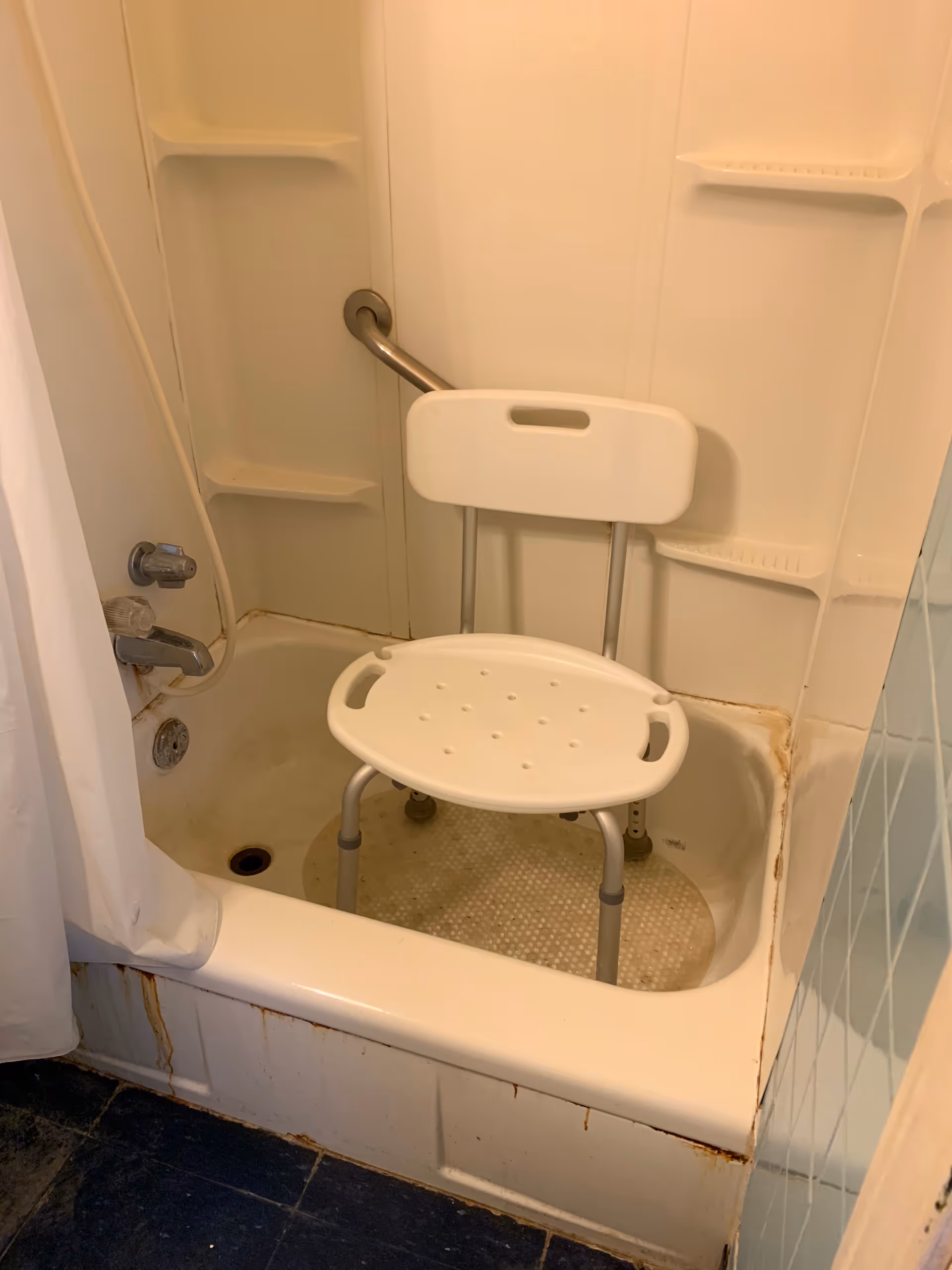 Shower stall in a bathroom with a white folding chair and grab bar.